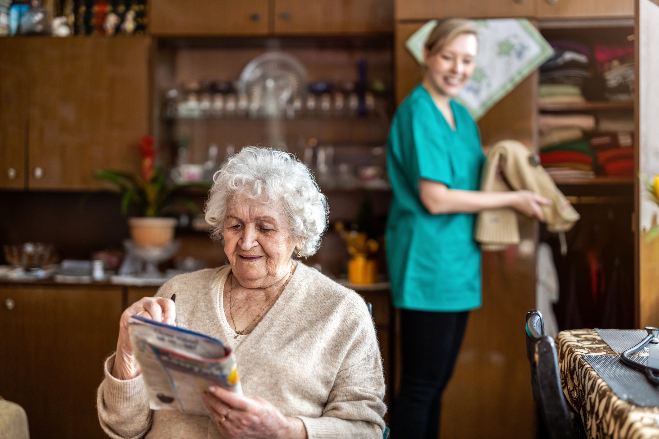 An elderly woman with white curly hair sitting at a table and looking at a package, with a caregiver in green scrubs standing in the background in a cozy kitchen.