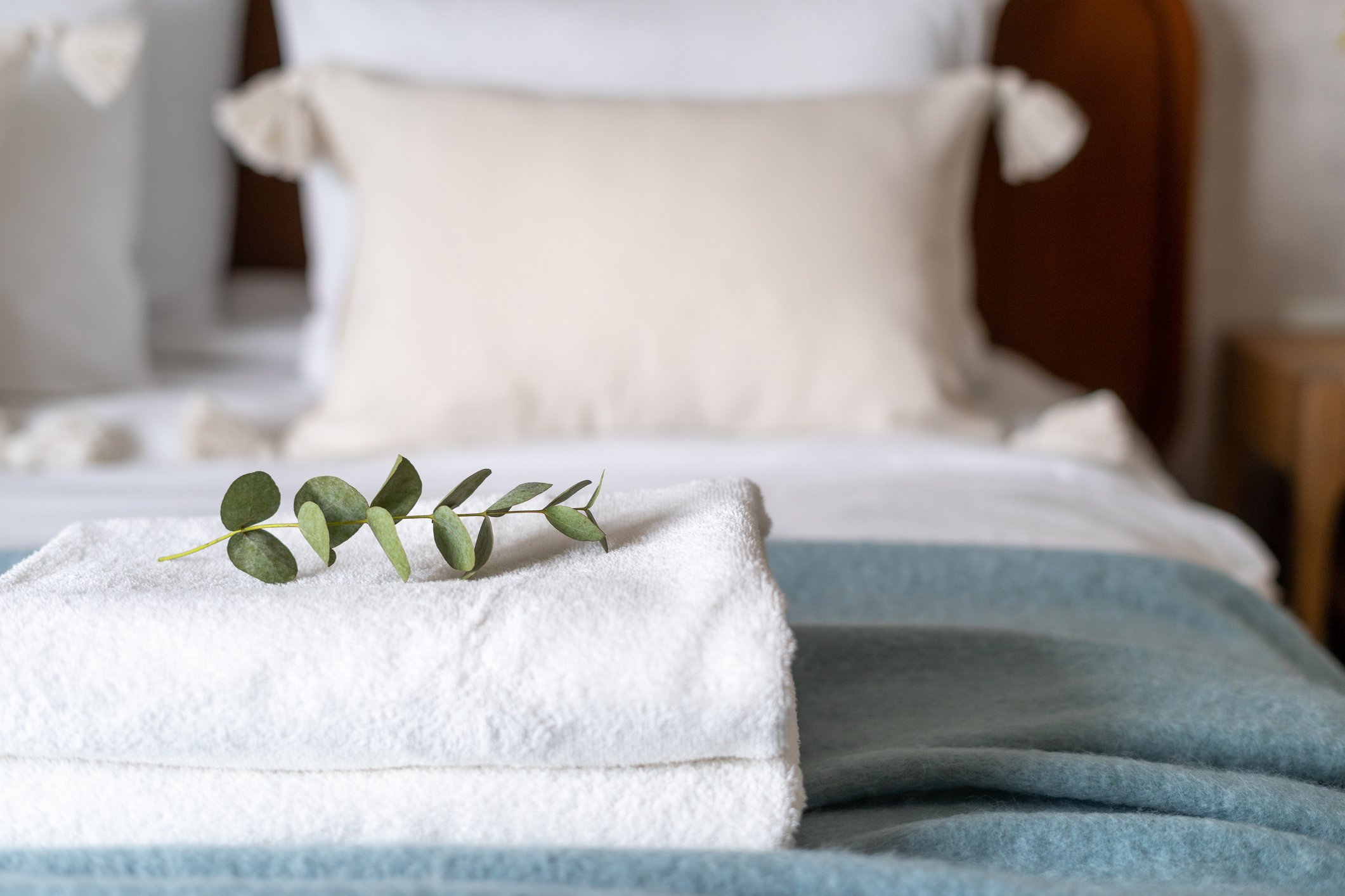 A neatly made bed with white sheets and a teal blanket, topped with folded white towels and a sprig of eucalyptus, in a cozy bedroom setting.