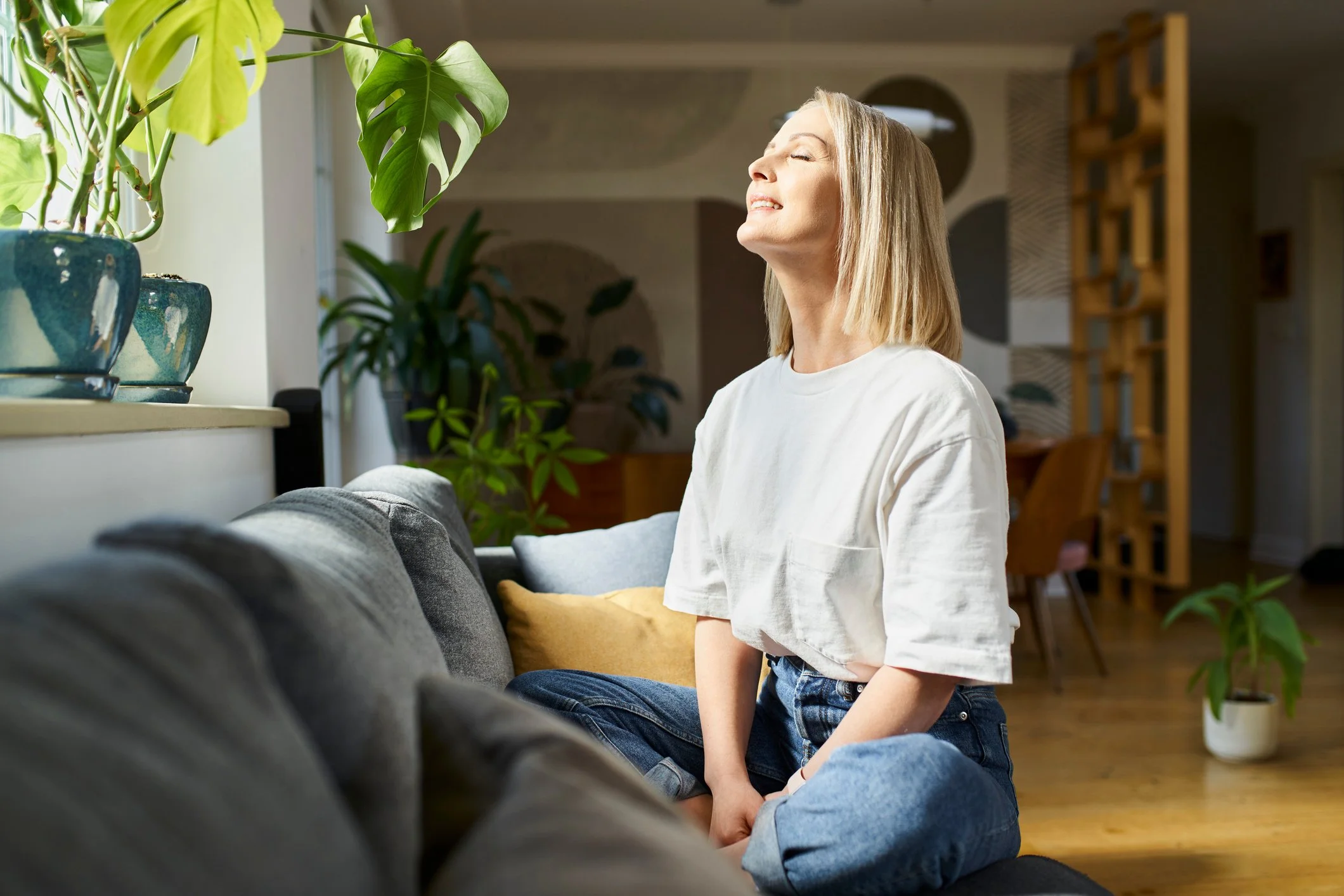 A woman with blonde hair sitting on a sofa with her eyes closed, enjoying natural sunlight from a window, in a cozy living room with plants and decorative items.