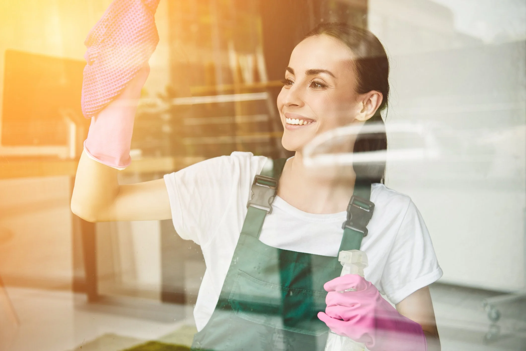 A woman cleaning a glass window with a spray bottle in her right hand and a pink cleaning cloth in her left hand, wearing pink rubber gloves, a white t-shirt, and a dark green apron, smiling and looking at the window.