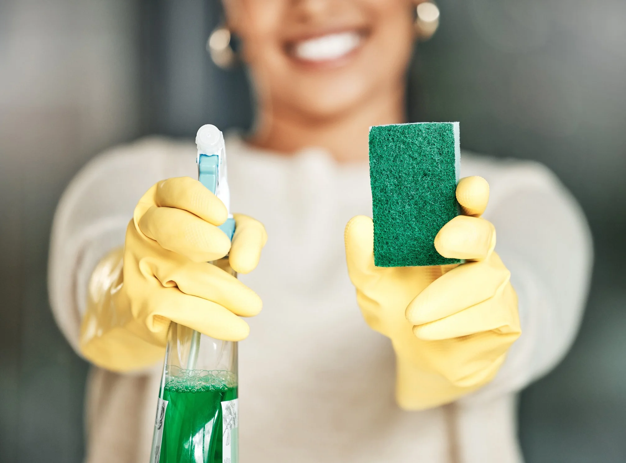 Person wearing yellow cleaning gloves holding a spray bottle with green cleaning solution and a green scrubbing sponge, smiling.