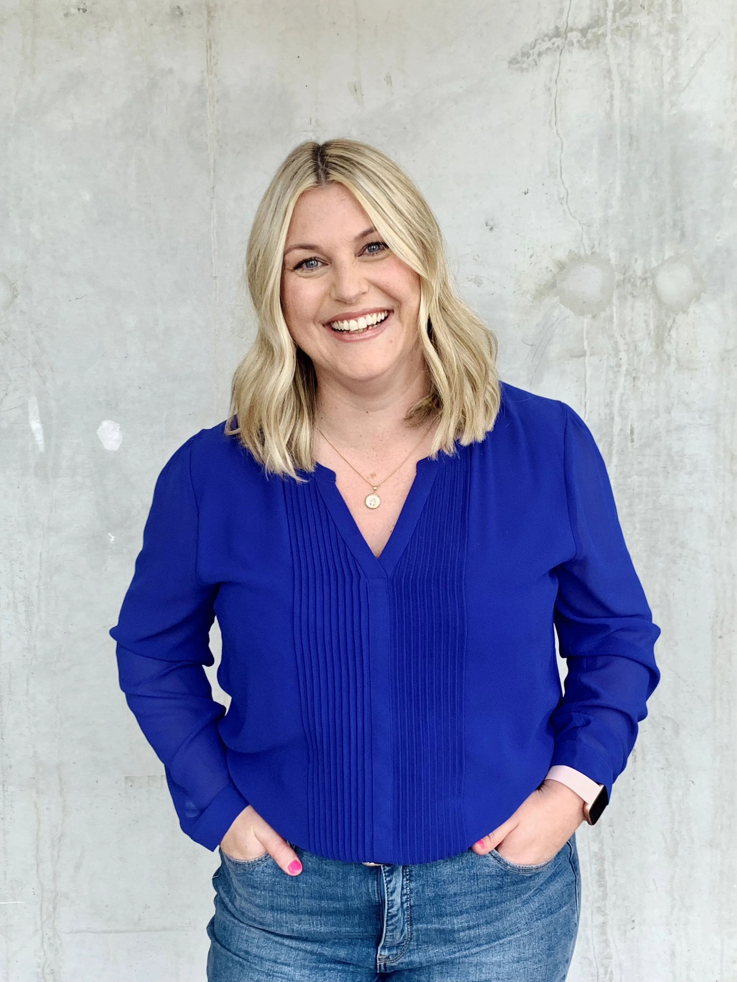 A woman with shoulder-length blonde hair, wearing a bright blue blouse and jeans, smiling at the camera in front of a plain gray wall.