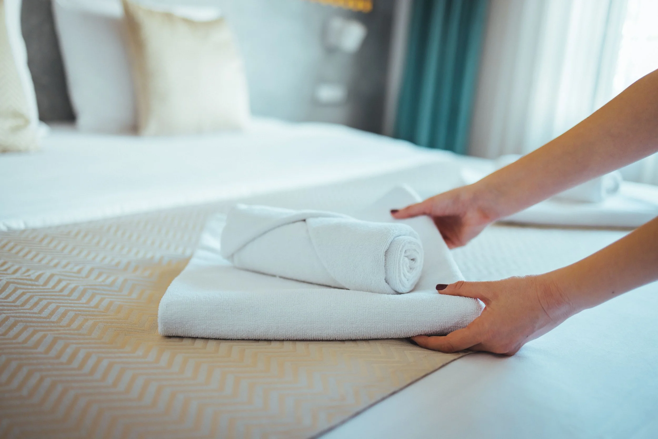 Person folding a white towel on a hotel bed with beige bed runner and pillows in the background