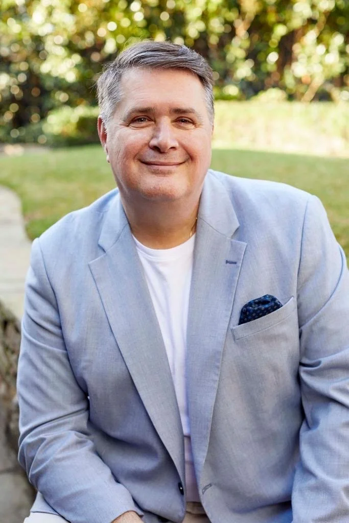 A smiling man wearing a light gray blazer and white t-shirt sitting outdoors with a blurred green background.