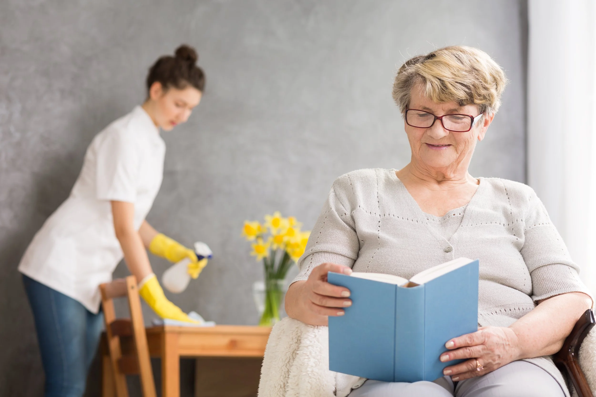 An elderly woman wearing glasses sits in a chair reading a book, while a young woman in a white shirt and yellow rubber gloves cleans a wooden table in the background.