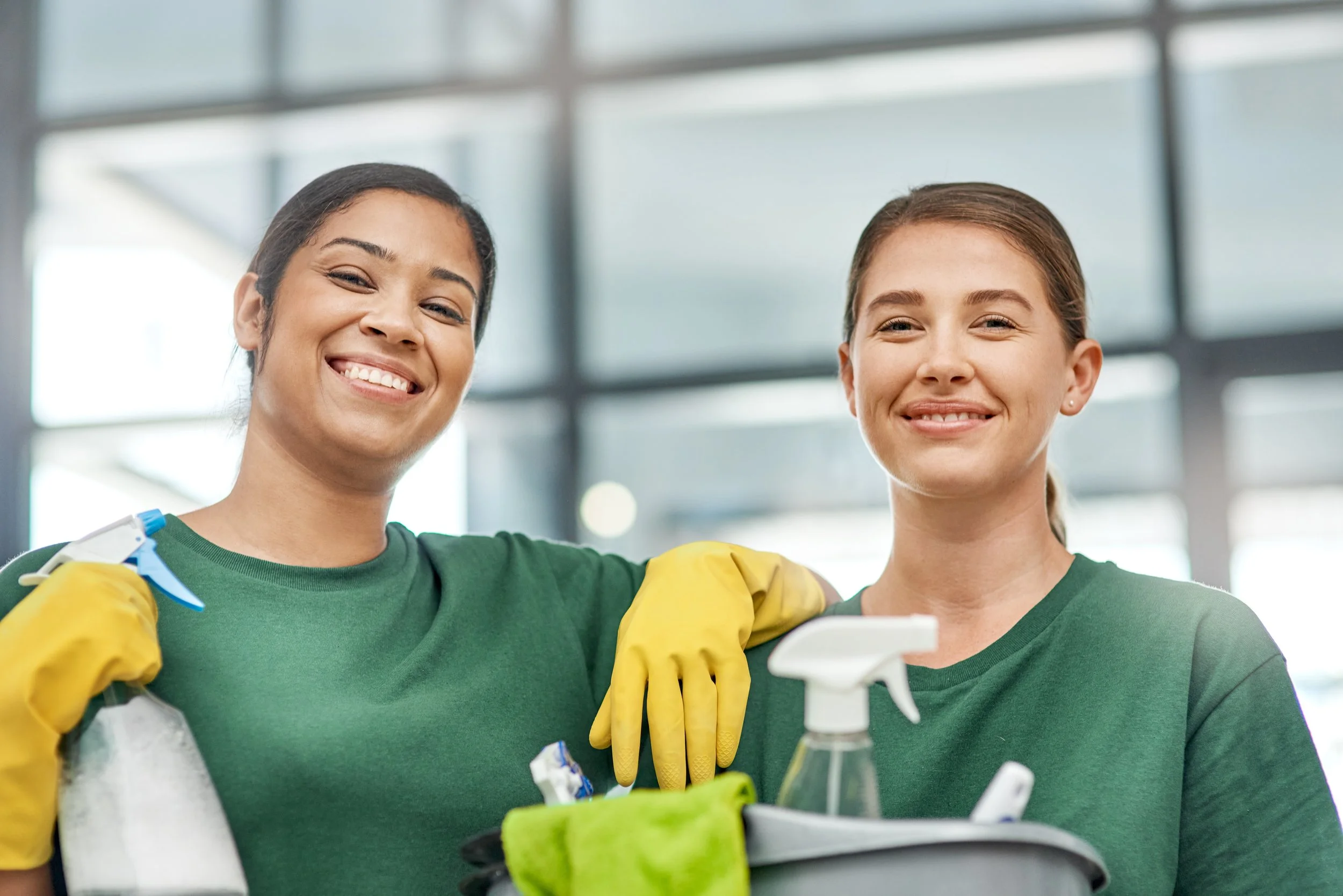 Two smiling women wearing green shirts and yellow gloves standing together with cleaning supplies, smiling at the camera.