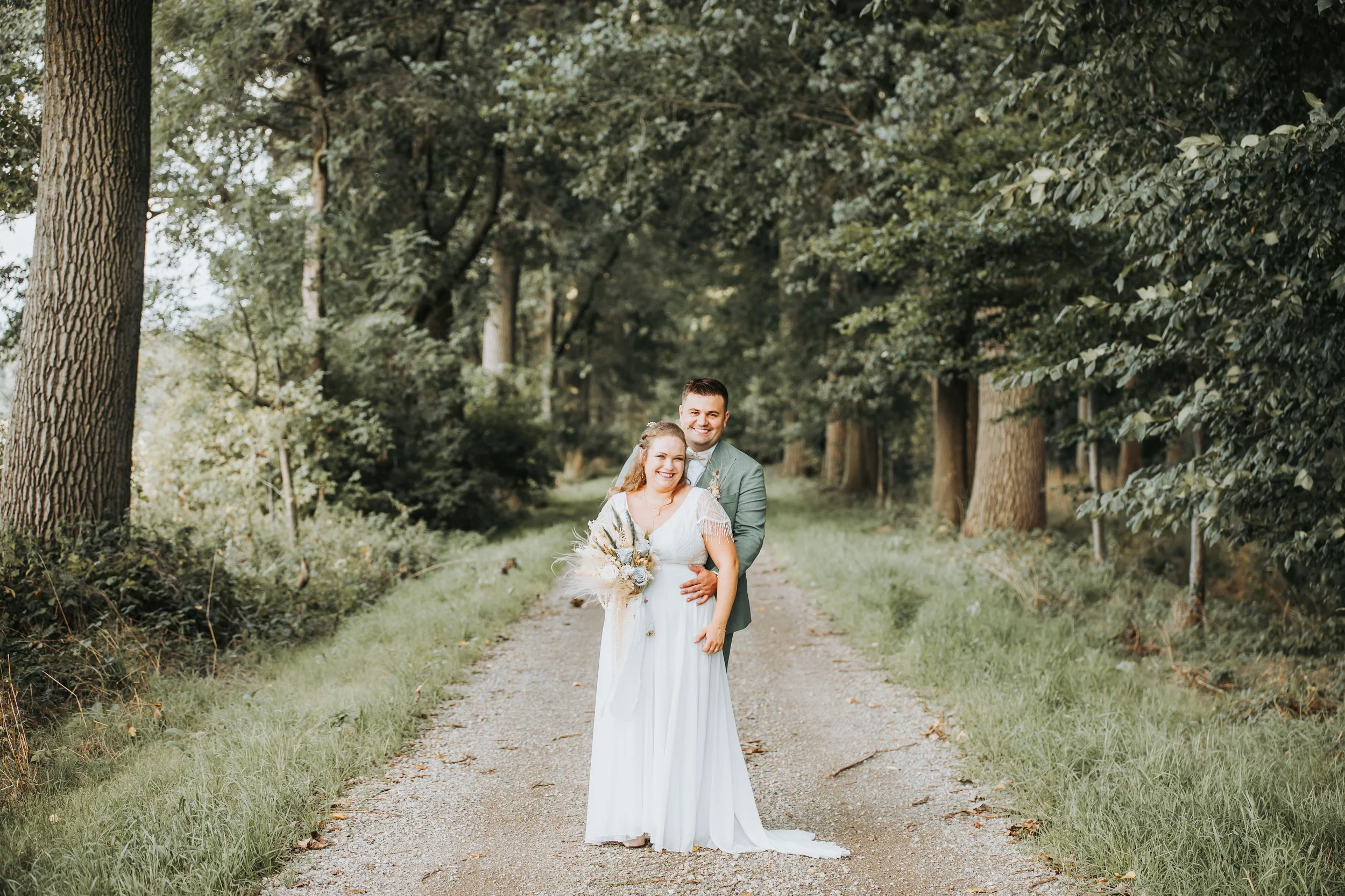 Een bruid en bruidegom op een bosweg, lachend en in trouwkleding, omringd door groene bomen.