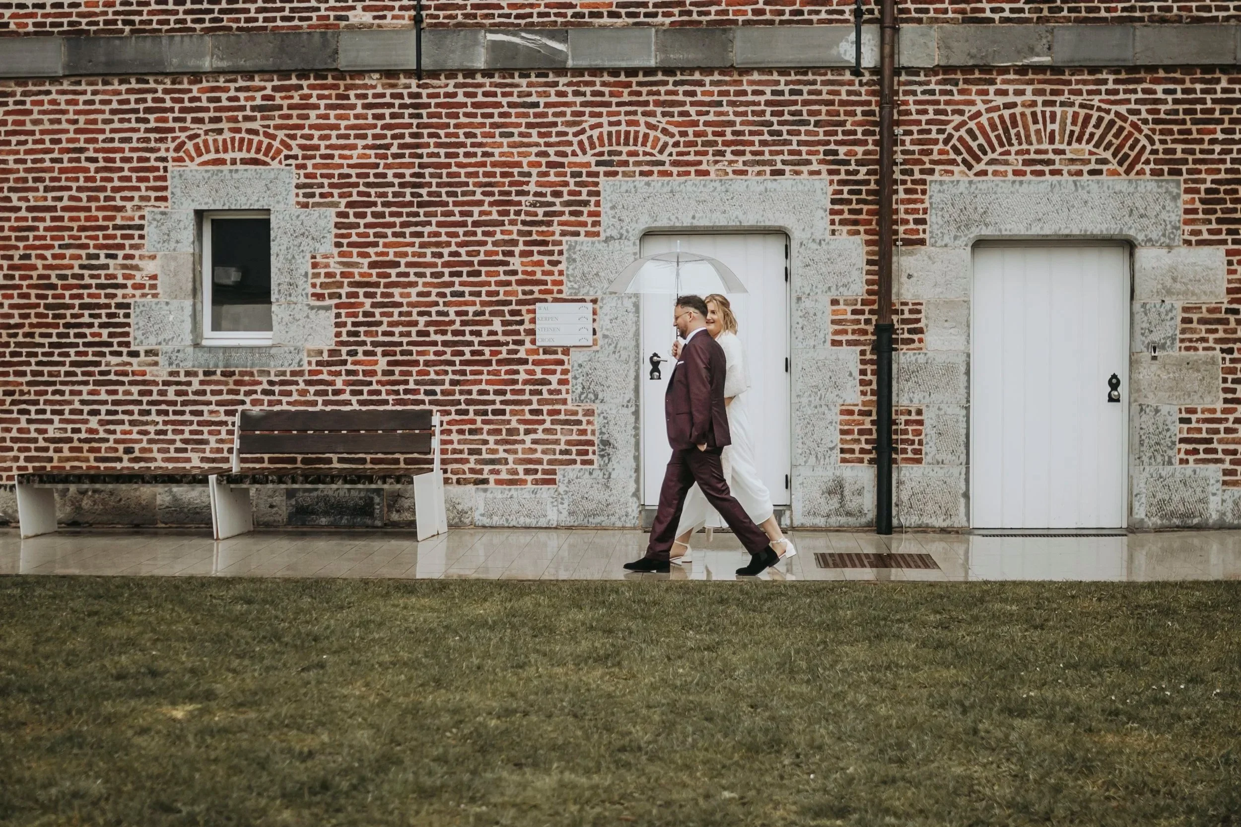 Twee mensen, een man en een vrouw, wandelen samen onder een doorzichtige paraplu op een natte stoep voor een bakstenen muur met witte en grijze details.