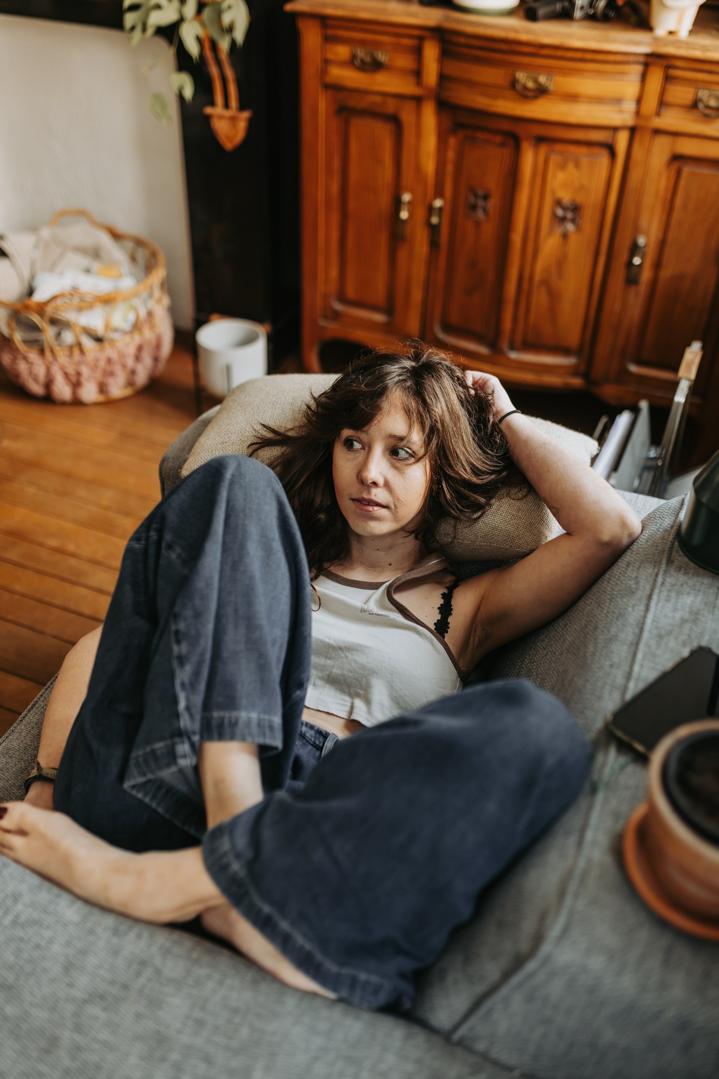 A young woman with brown hair is lying on a beige couch, resting her head on a throw pillow, with her arm behind her head, looking slightly to her left.