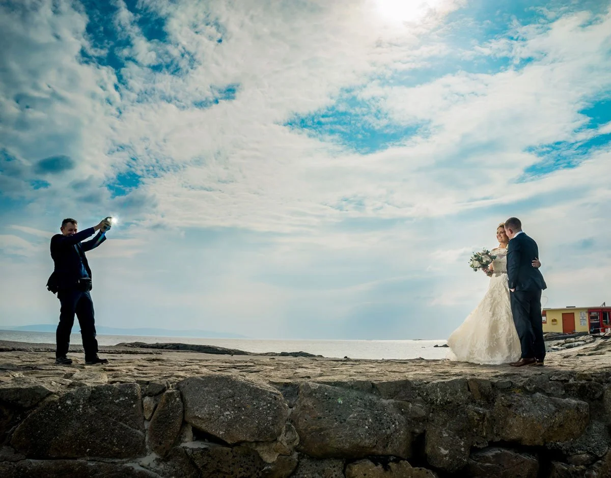 A bride and groom standing on a stone ledge at the beach, with a person taking their photo against a cloudy sky and ocean background.