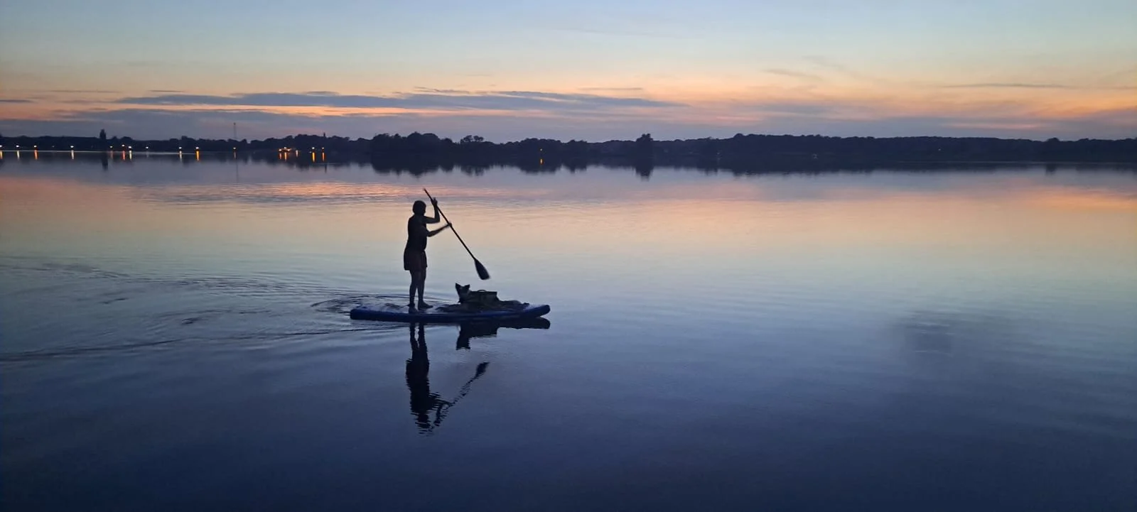 Person auf einem SUP-Board mit Hund auf ruhigem Wasser bei Sonnenuntergang, die Silhouetten sind sichtbar