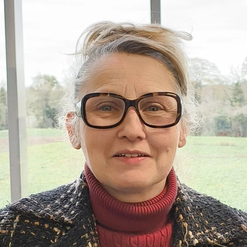 Illustration of a young woman with blonde hair wearing glasses and a patterned orange and white kimono-style top, standing in a corridor with light-colored walls and blue flooring.