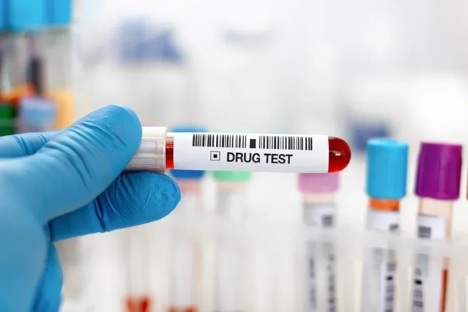 Gloved hand holding a blood sample vial labelled Drug Test in front of a rack of colourful laboratory tubes