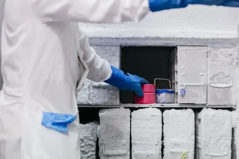 Scientist in white lab coat and blue gloves retrieving a sample from a heavily frosted ultra-low temperature laboratory freezer