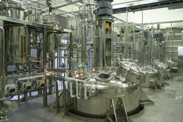 Large-scale industrial bioreactor facility with multiple stainless steel fermentation tanks, pipes, and processing equipment arranged in rows inside a pharmaceutical or biotech manufacturing plant