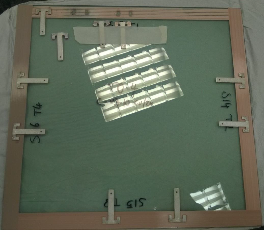 A glass picture frame with small wooden L-shaped corner brackets, a green background, and handwritten measurements and notes. The reflection of overhead lights is visible on the glass surface.