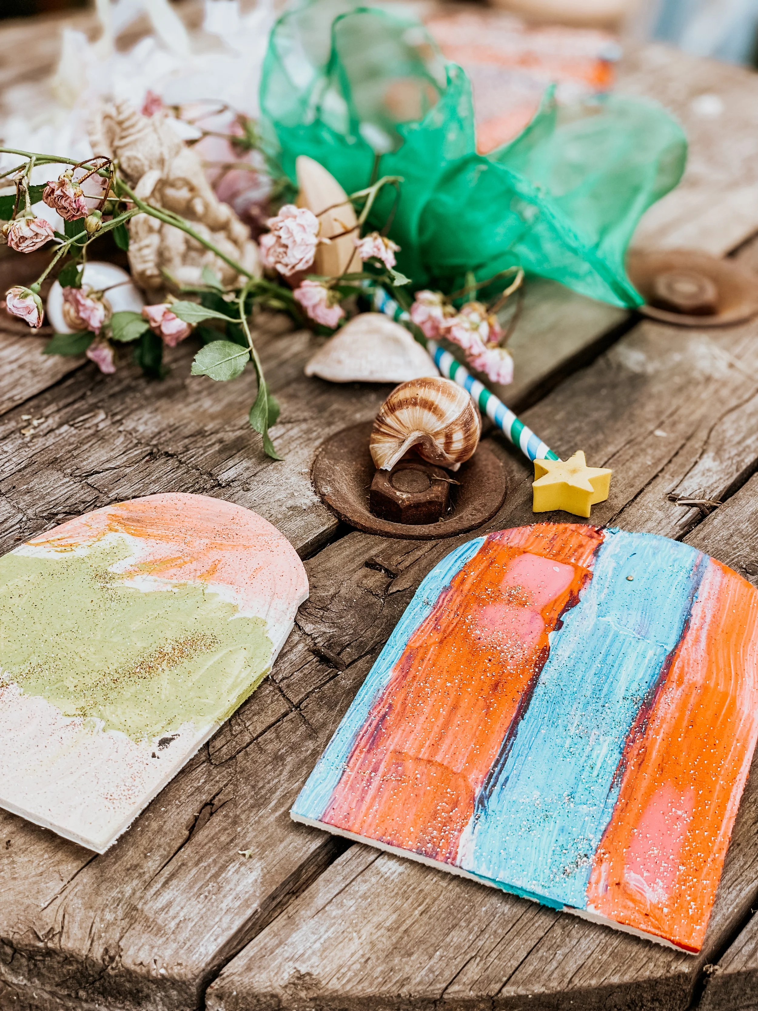 Bunte, abstrakte Gemälde auf Holzuntergrund, umgeben von Muscheln, Blumen, einer grünen Schleife, einem Stern aus Knete und einem Papierstrohhalm.