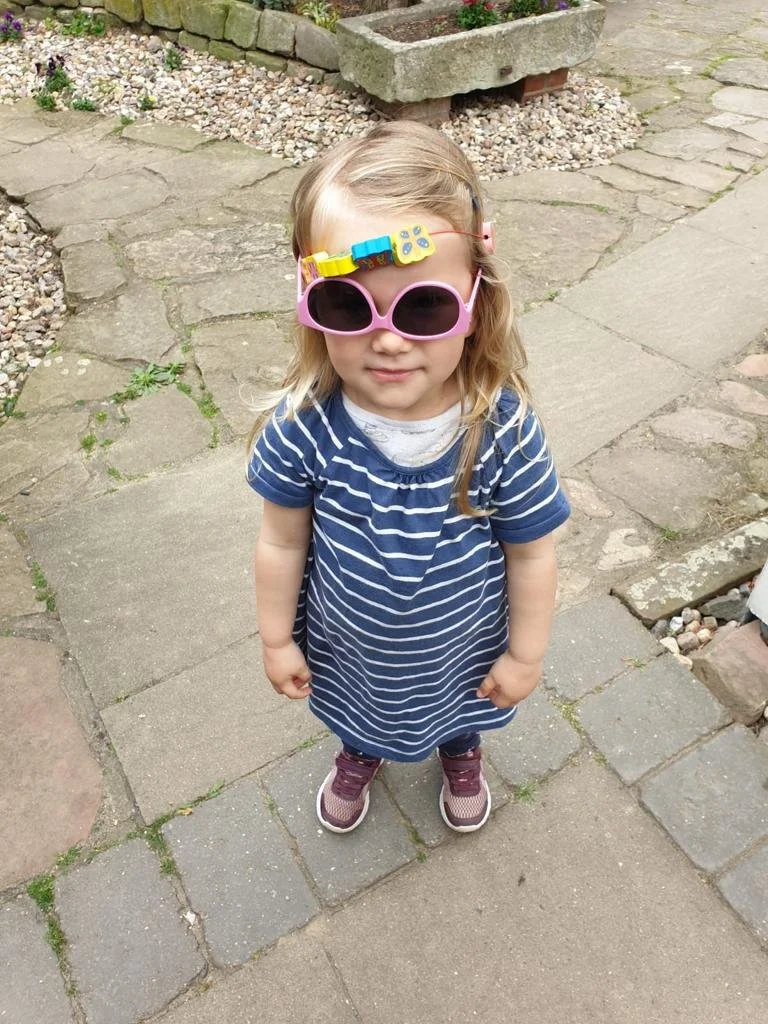 Ein kleines Mädchen steht auf einem gepflasterten Weg, trägt eine blau-weiße gestreifte Kleider und große rosa Sonnenbrille, sowie eine Kindersauna und Sneakers.