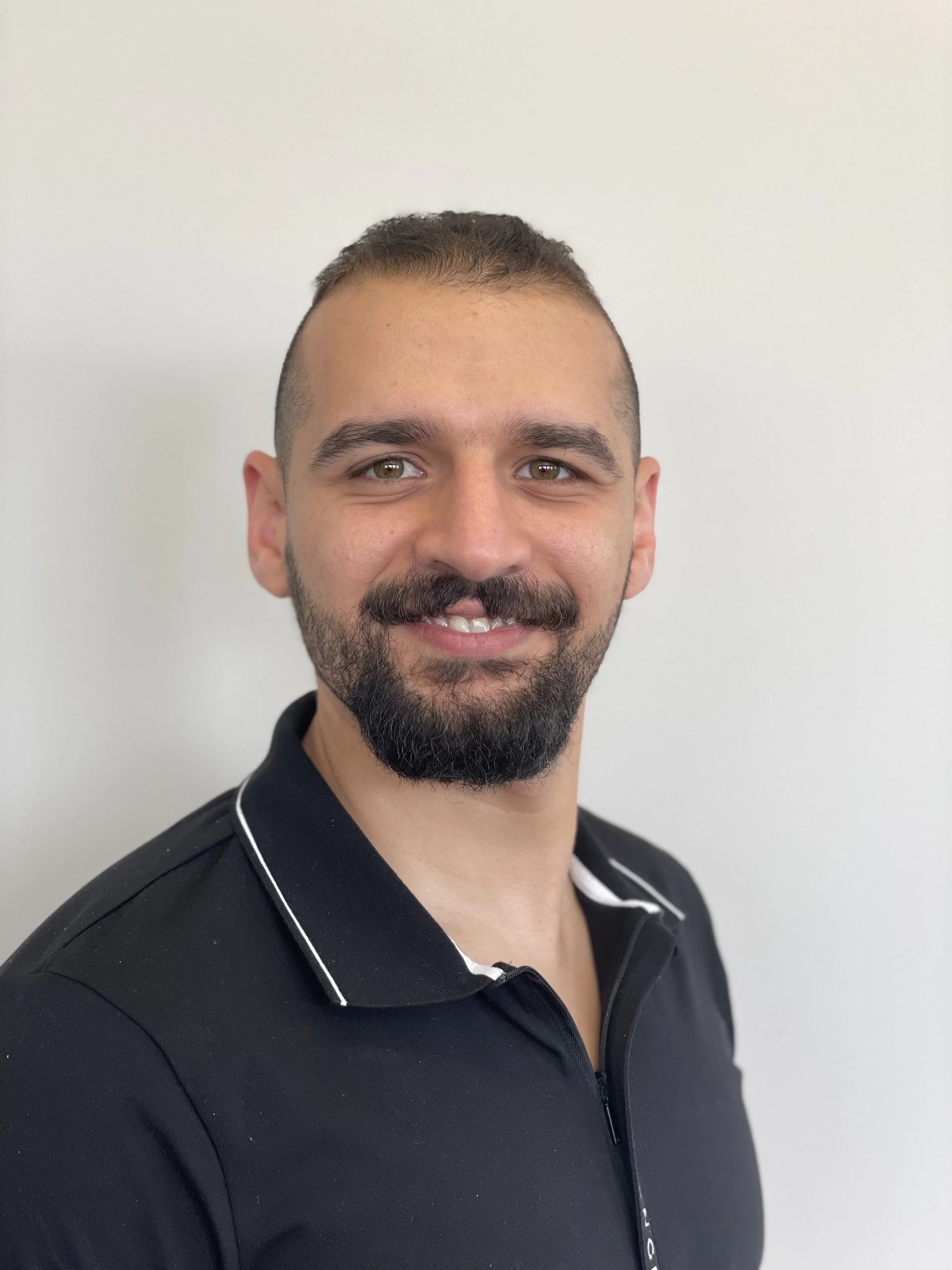 A smiling man with short dark hair, a beard, and a mustache, wearing a black polo shirt with white trim, standing against a plain white background.