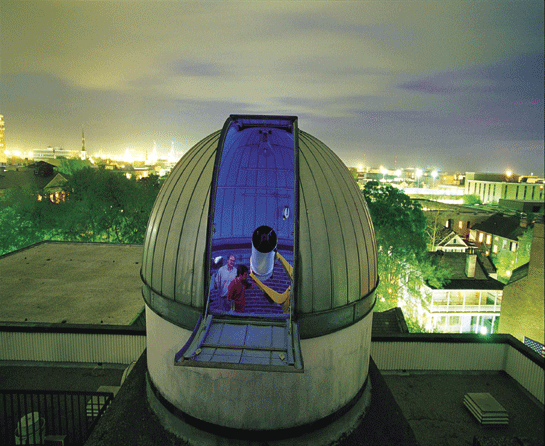 View from the top of an observatory dome with a large telescope and two people taking a selfie at night, with city lights in the background.