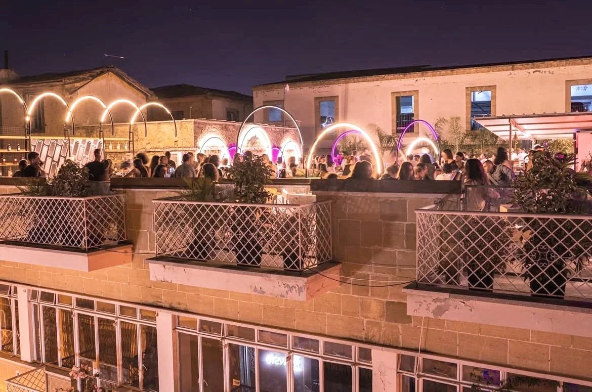 People gathering on a rooftop terrace at night with illuminated arches and city buildings in the background.