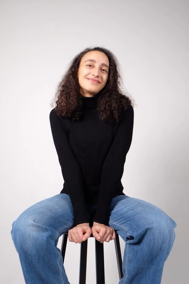A young woman with curly brown hair, sitting on a black stool, smiling, wearing a black turtleneck sweater and blue jeans, against a plain white background.