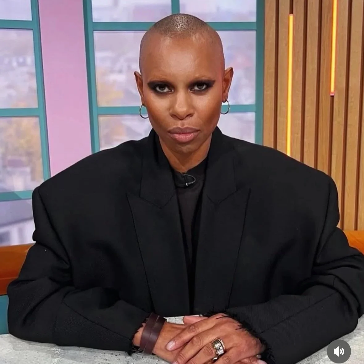 A person with a shaved head sitting at a table, wearing a black blazer, with intense eye makeup and hoop earrings, looking directly at the camera in a brightly colored room.