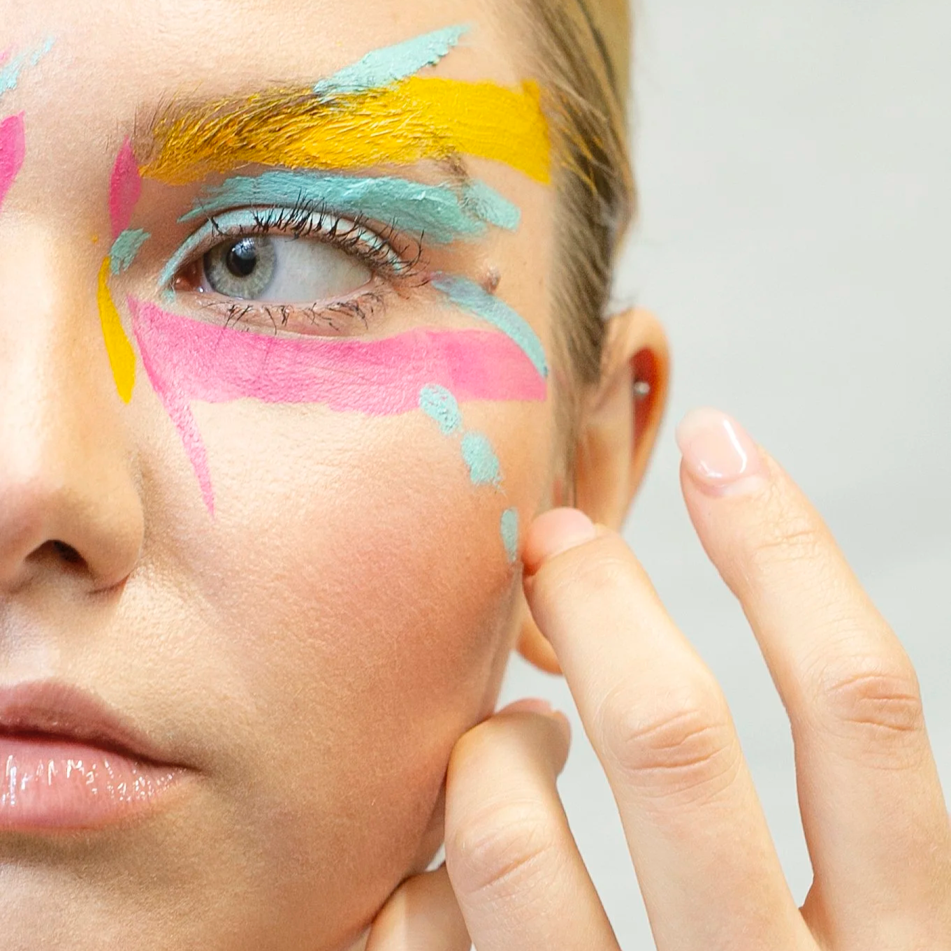 Close-up of a woman's face with colorful abstract makeup around her eye, including pink, blue, and yellow paint strokes, and her hand touching her cheek.