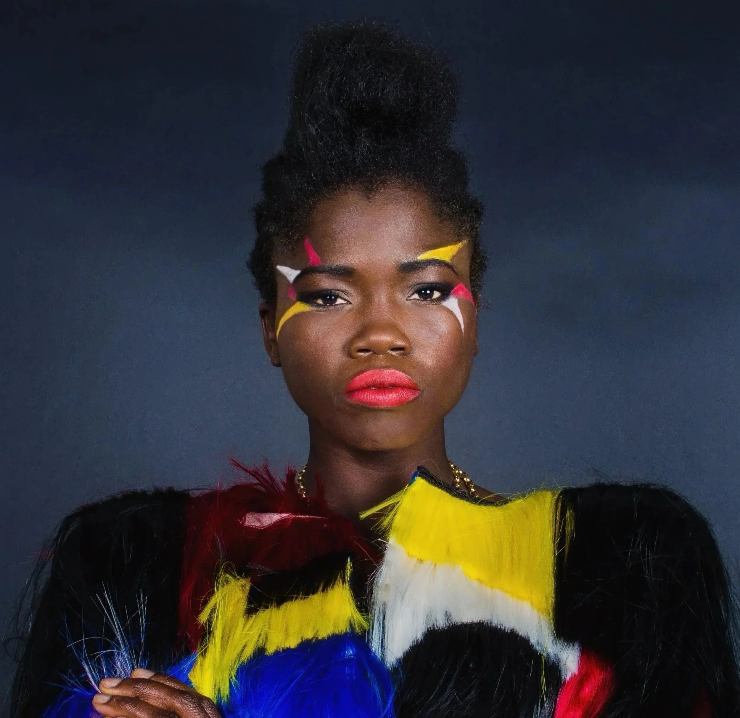 A woman with dark skin wearing colorful feathered clothing and face paint against a dark background.