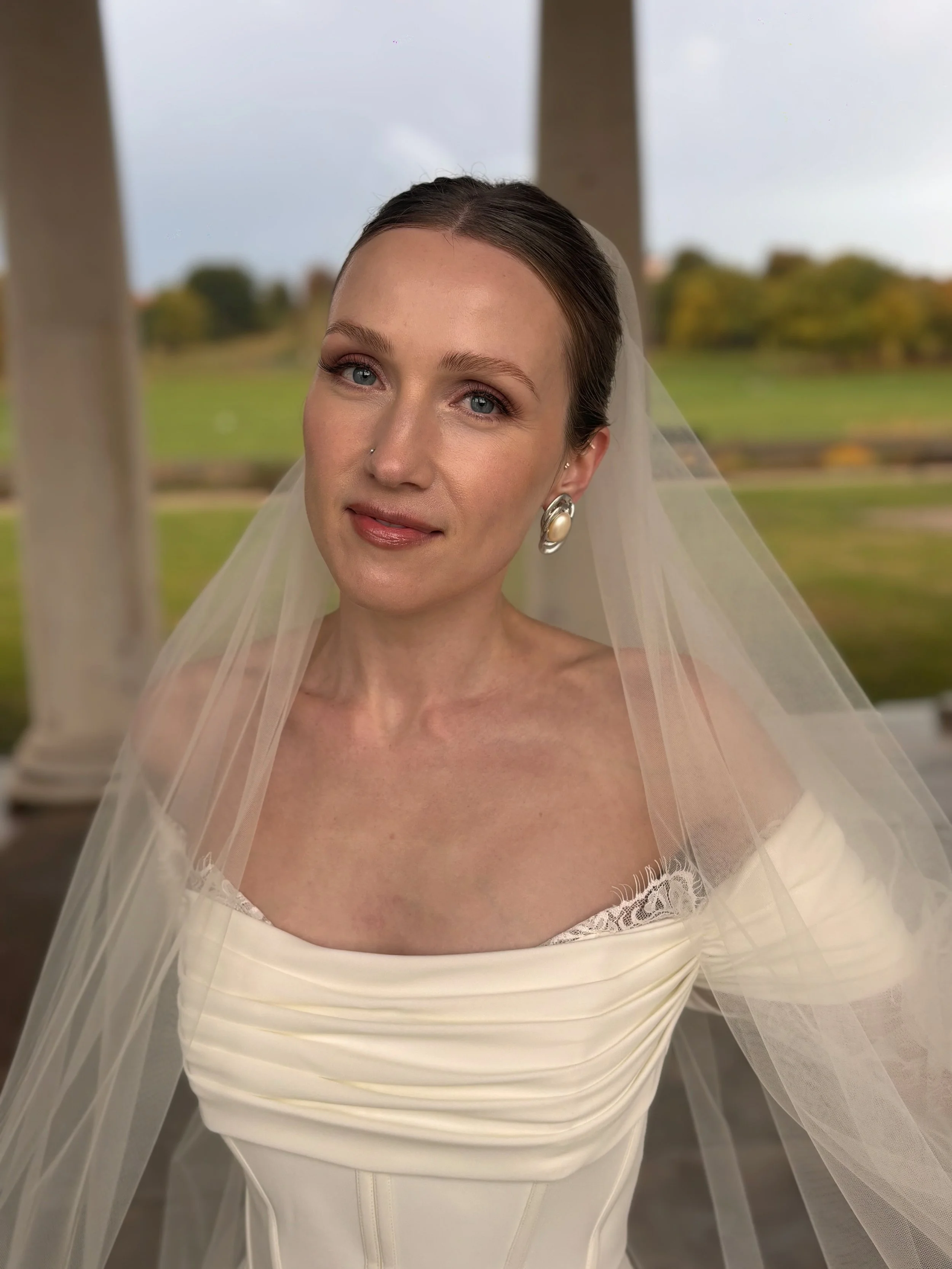 A woman in a wedding dress with a veil, standing outdoors with green landscape and cloudy sky in the background.