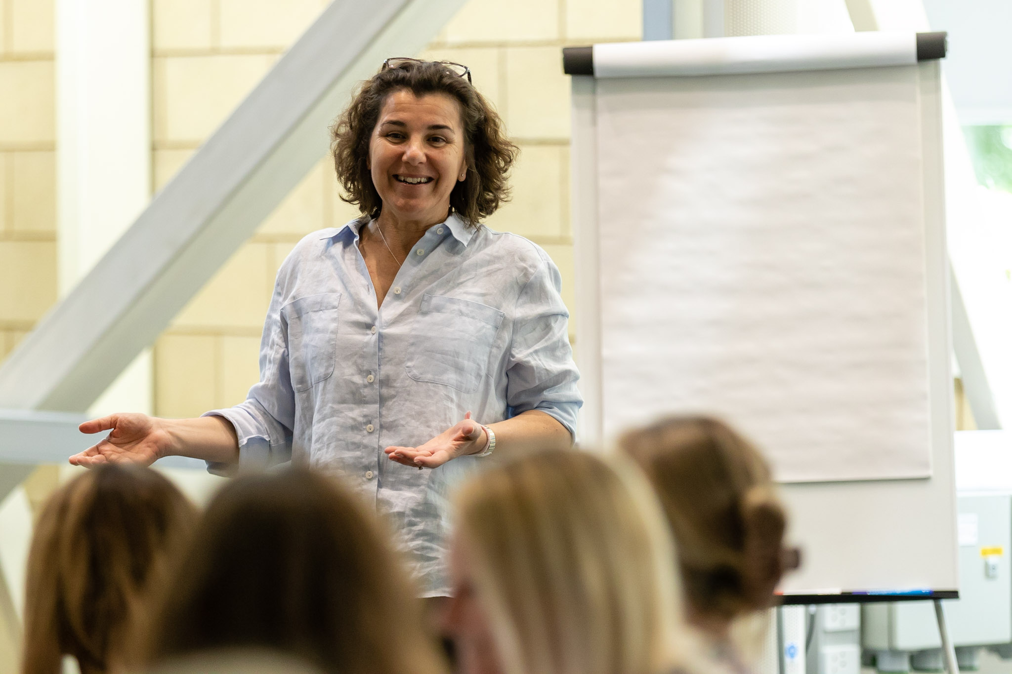 A woman giving a presentation in front of an audience, standing next to a blank flip chart, with her hands open.