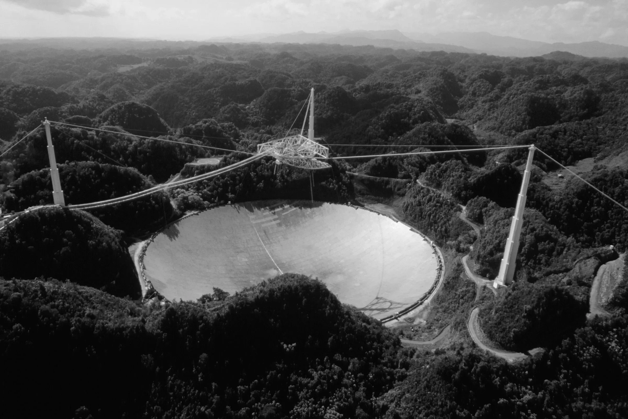 Black and white aerial photo of a large radio telescope dish surrounded by forested hills with winding roads leading to the structure.