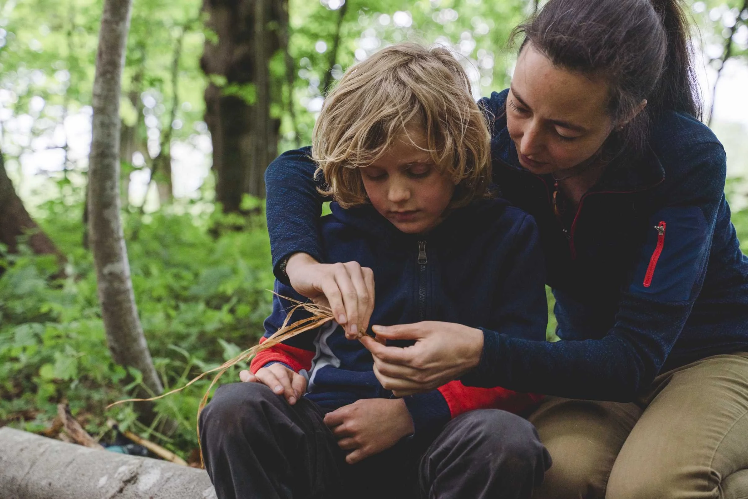 Eine Frau zeigt einem Jungen, wie man eine Schnur aus Naturmaterialien dreht im Wald, beide sitzen auf einem Baumstamm.