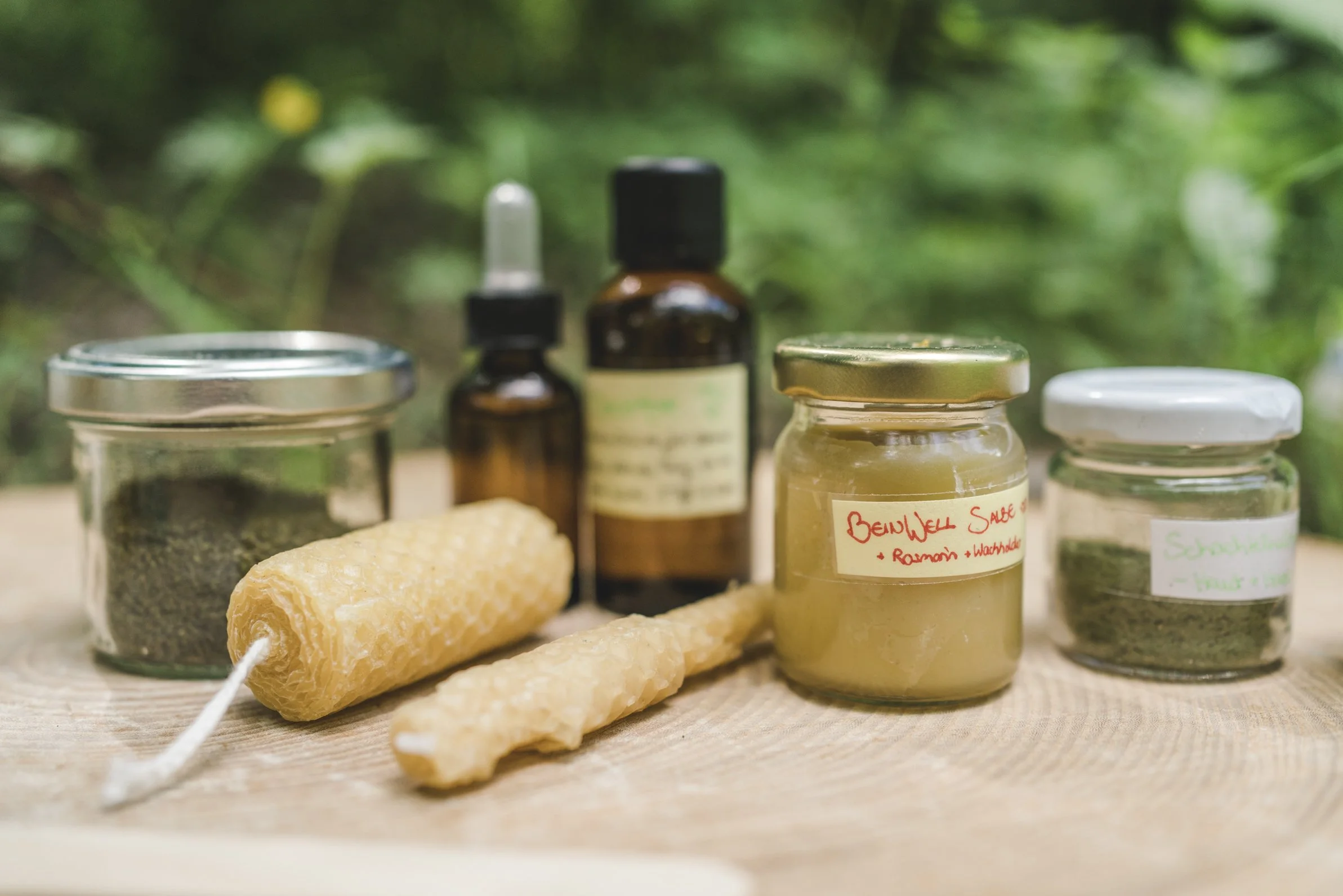 Natürliche Heilmittel und Kräuter in Gläsern, Fläschchen und Wachsblock auf Holzuntergrund mit grüner, unscharfer Natur im Hintergrund.