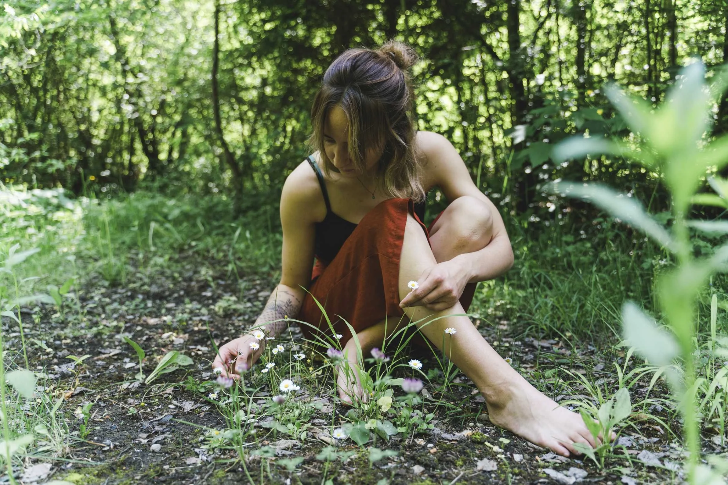Eine Frau sitzt barfuß im Wald, pflückt kleine weiße Blumen.