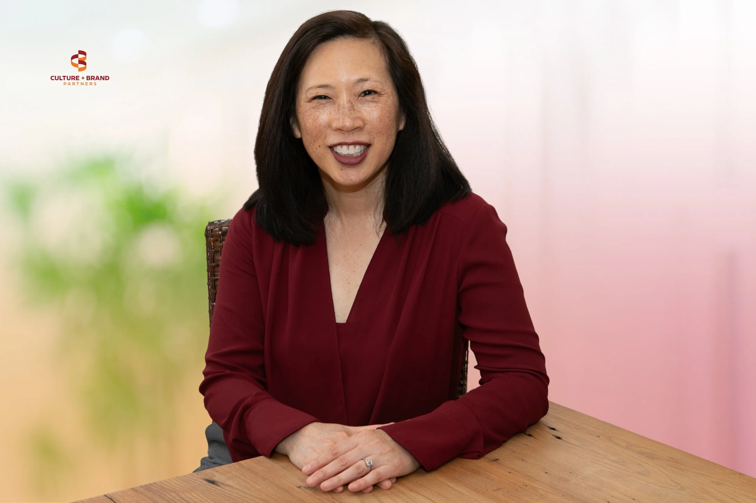 A woman, Serena Fong, with shoulder-length black hair, wearing a red blouse, smiling and sitting at a wooden table.