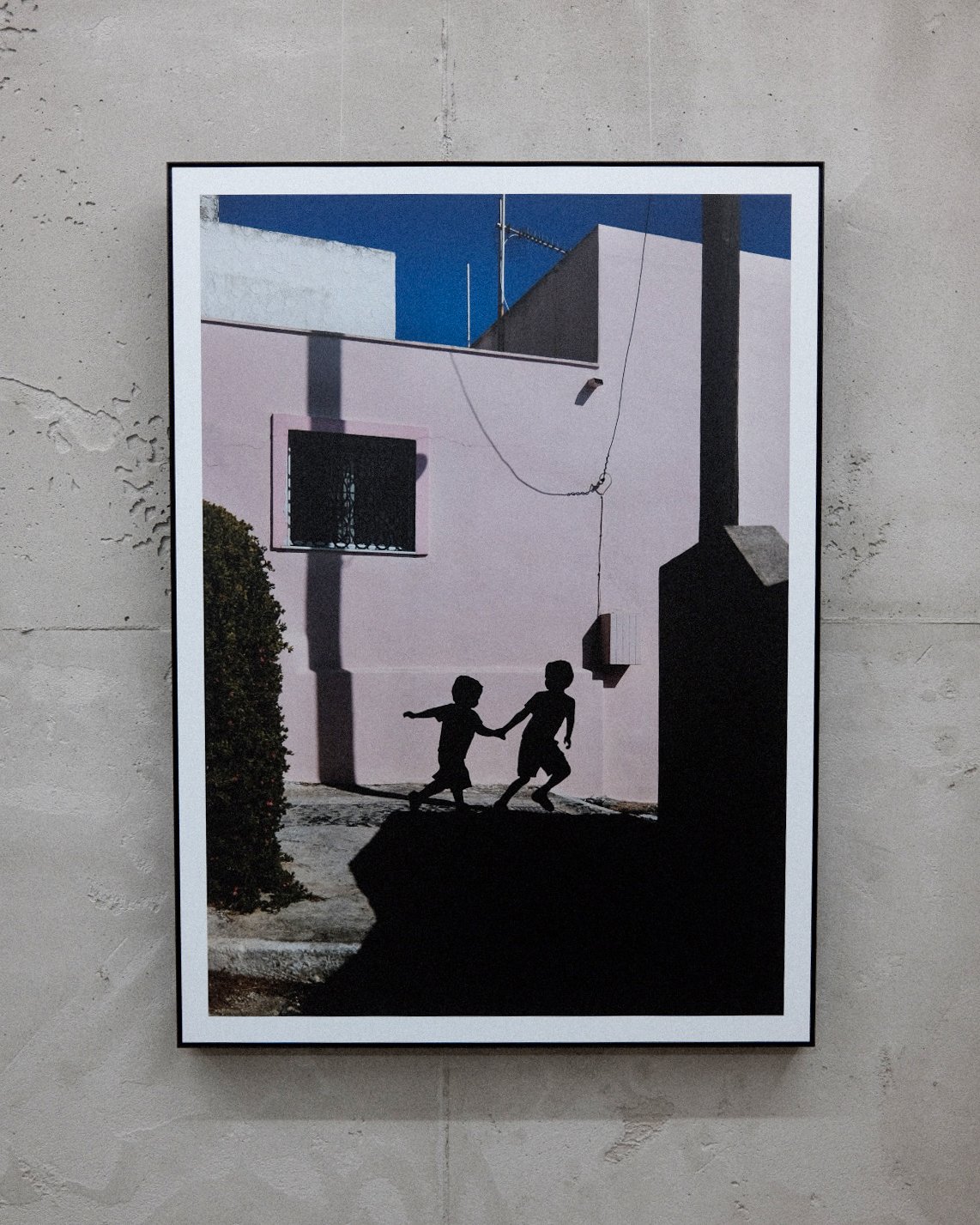 Schatten von zwei Kindern, die sich an einer Wand im Schatten halten, vor einer rosa Wand mit Fenster und Schädelmuster, bei sonnigem Himmel.