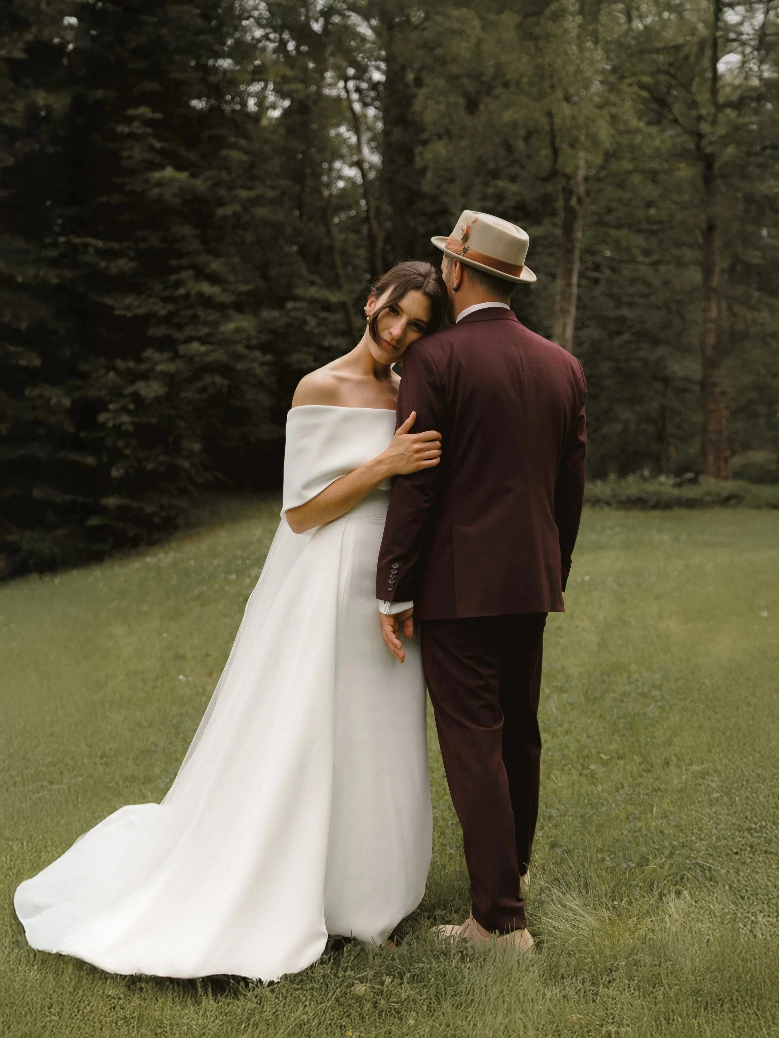 Ein Paar in Hochzeitskleidung steht auf einer Wiese in einem Wald, die Frau trägt ein weißes Hochzeitskleid und der Mann einen dunklen Anzug mit Hut.