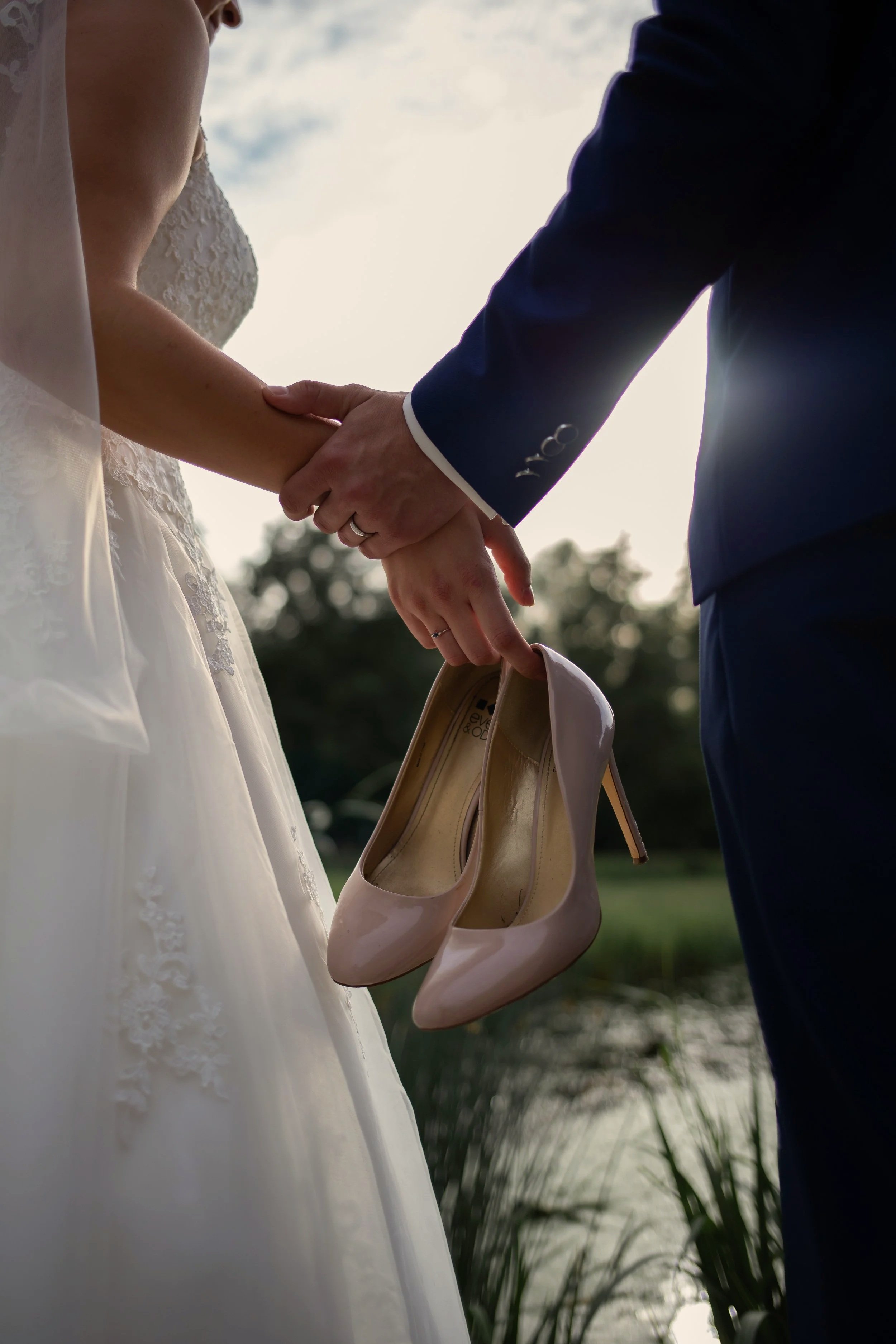 Ein Brautpaar hält sich die Hände bei ihrer Hochzeit, die Braut trägt ein weißes Brautkleid, der Bräutigam trägt einen dunklen Anzug, und die Brautschuh ist sichtbar, während sie im Freien bei Sonnenuntergang stehen.