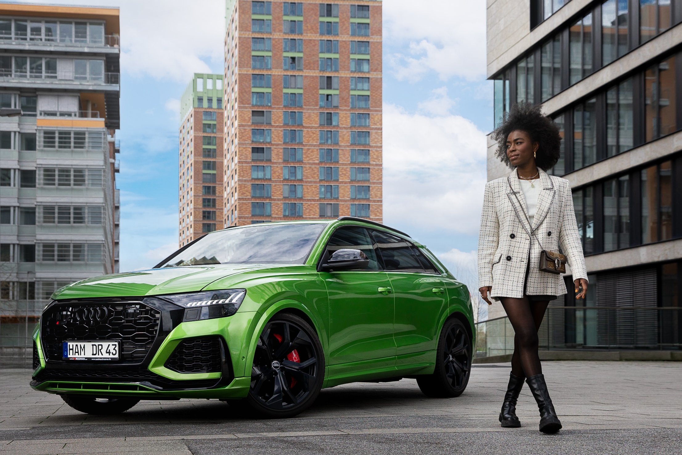 Einelyh stämmige frau steht neben einem grünen Audi e-tron in einer urbanen Umgebung mit Hochhäusern im Hintergrund. Sie trägt einen weißen karierten Blazer, schwarze Schuhe und trägt eine kleine Umhängetasche.