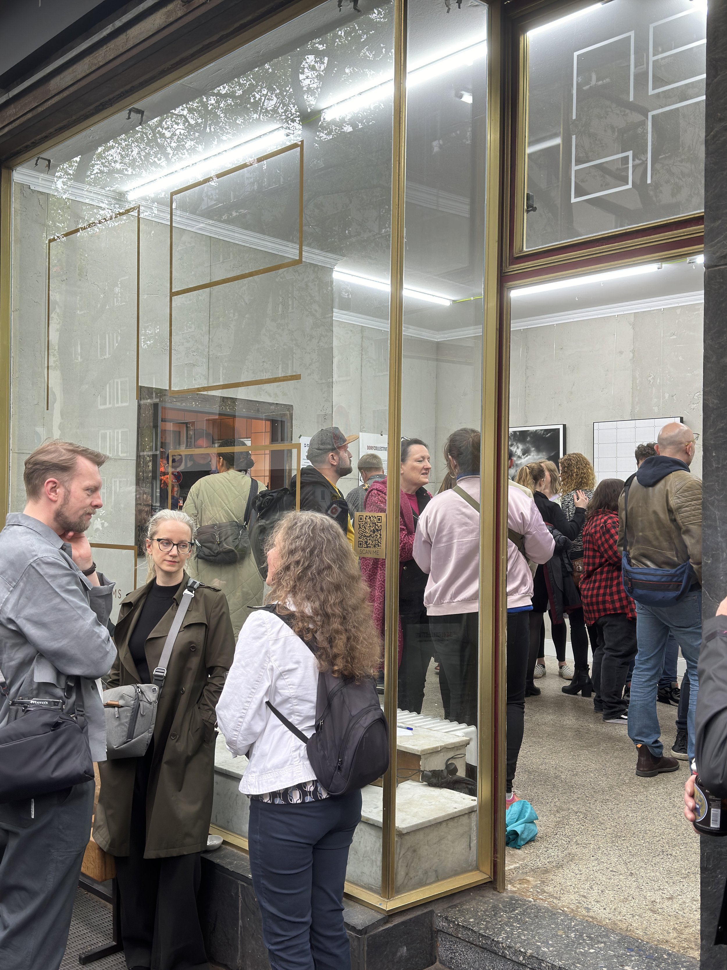Menschenmenge und Warteschlange vor einer Galerie oder Ausstellung, sichtbar durch die Glasscheibe. Einige Leute sprechen miteinander, andere schauen sich um.