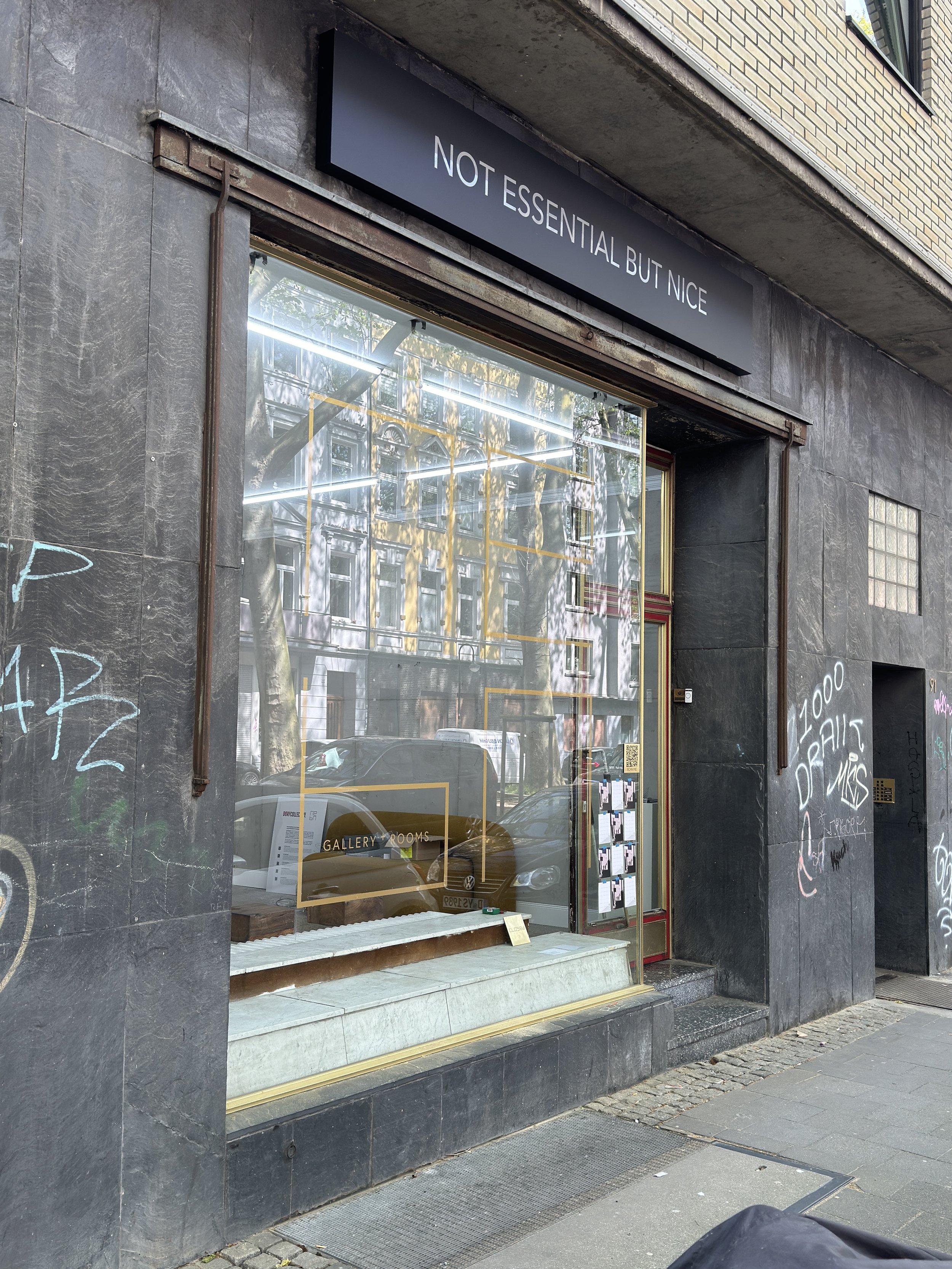 Schaufenster eines Galerieraums mit dem Schild 'NOT ESSENTIAL BUT NICE' darüber. Das Fenster spiegelt ein Gebäude und Autos wider, während Graffiti die schwarze Wand umgibt.