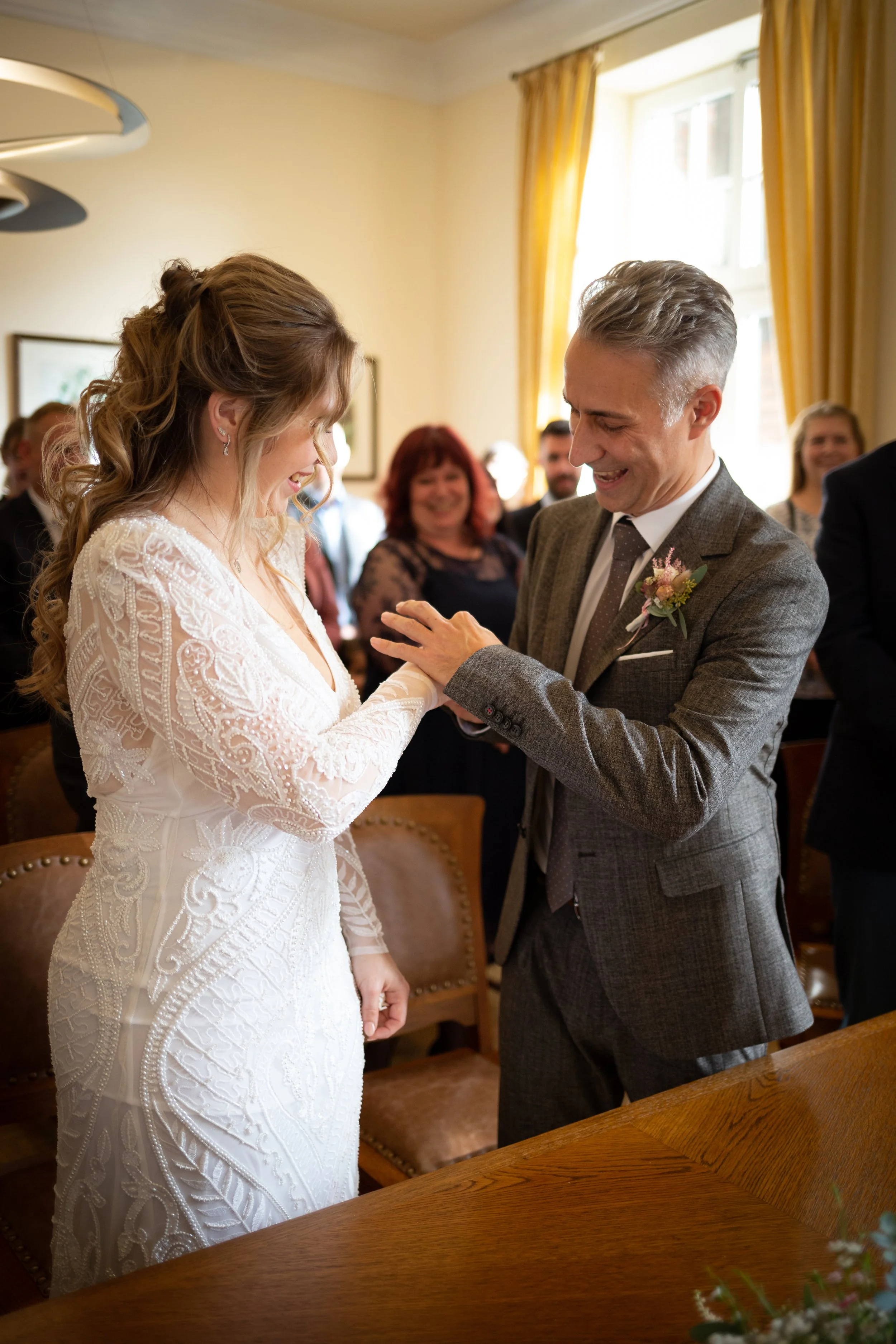 Ein Bräutigam in Anzug berührt die Hand einer Braut in einem Hochzeitskleid bei ihrer Trauung, umgeben von lächelnden Gästen in einem festlich dekorierten Raum.