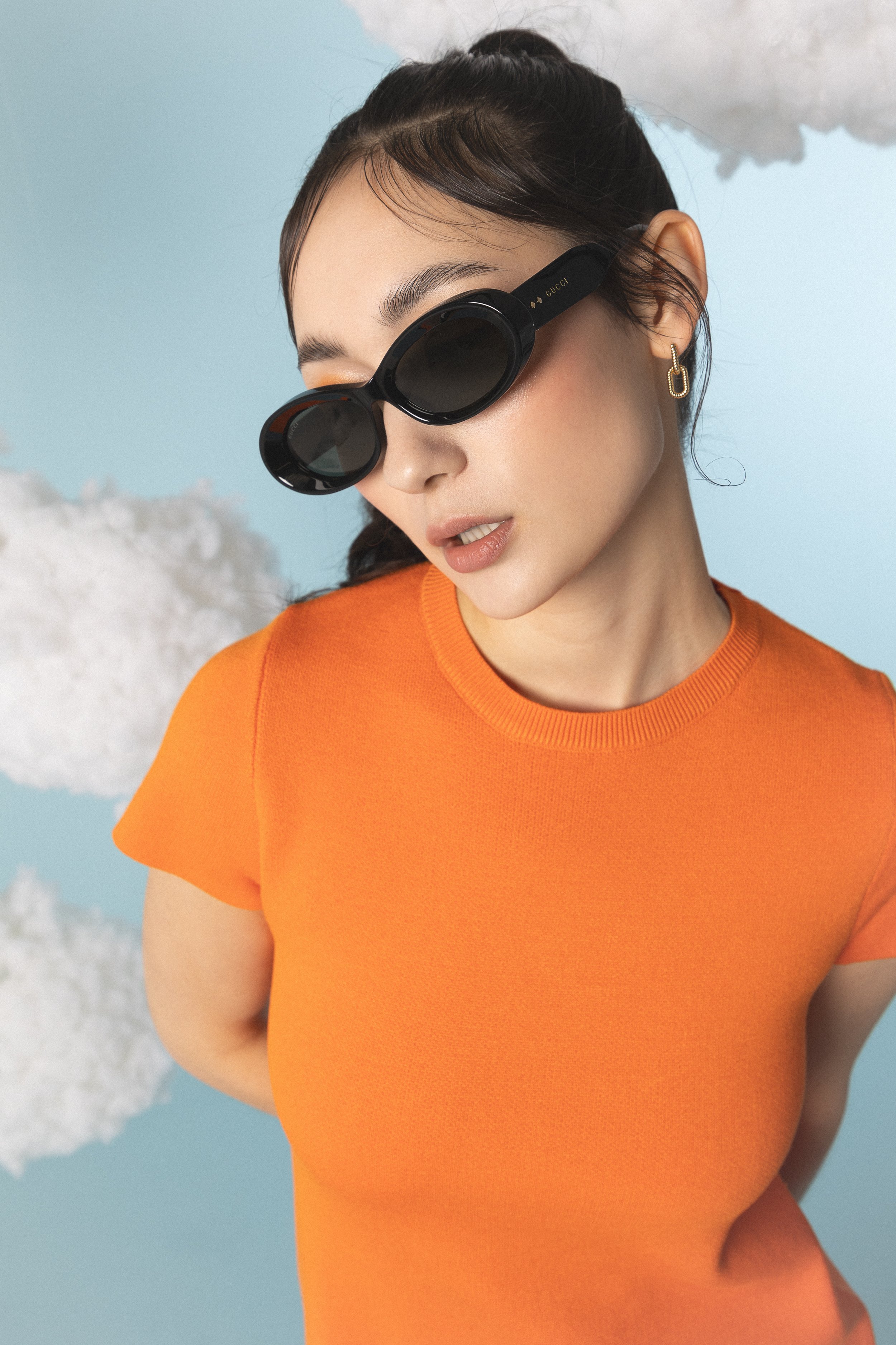 Eine junge Frau trägt große schwarze Sonnenbrillen und ein orangefarbenes T-Shirt, mit einem Himmel mit Wolken im Hintergrund.