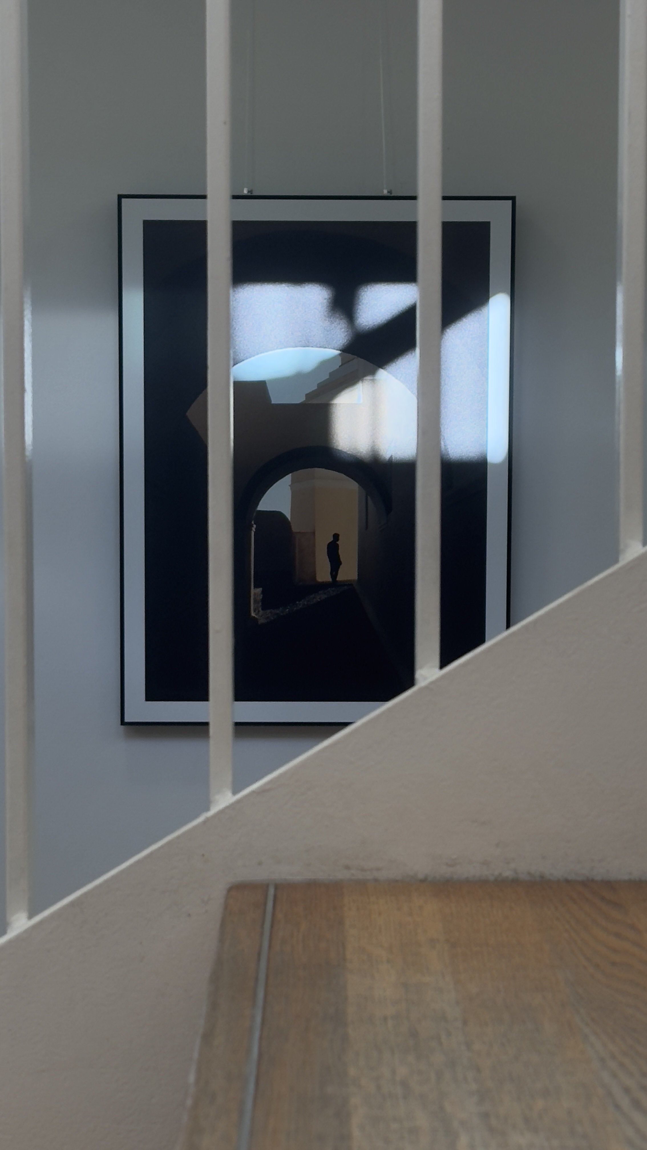 Fotografie eines Kunstwerks mit einer Silhouette eines Menschen, aufgenommen durch einen Geländer, mit Licht und Schatteneffekten.