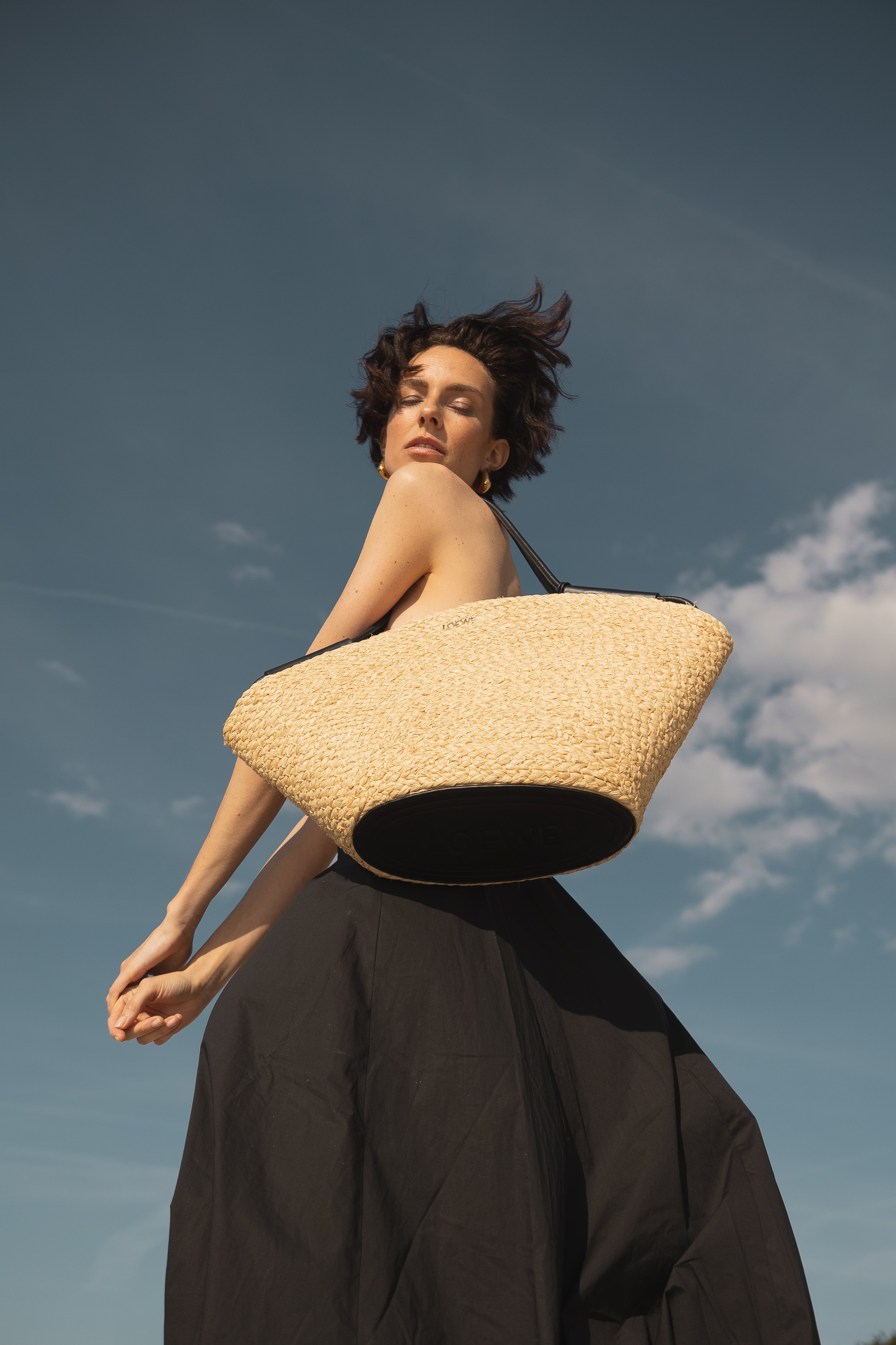 Frau mit braunen, kurzen, windbewegten Haaren trägt eine große, geflochtene Sonnenbrille und eine schwarze Hose, während sie eine große Strandtasche trägt. Der Himmel im Hintergrund ist blau mit einigen Wolken.
