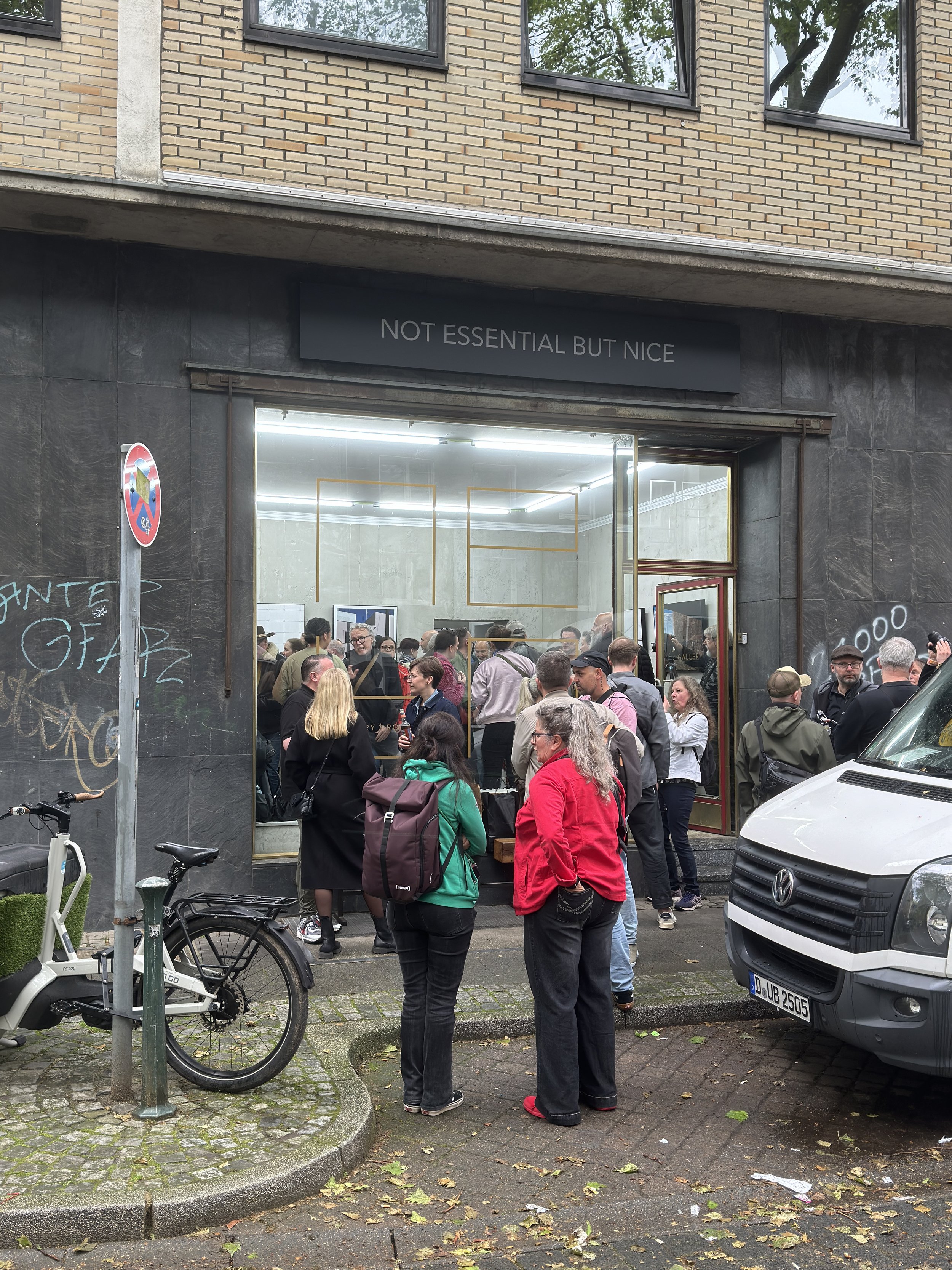 Menschenmenge vor einem Kunstgalerie-Eingang mit dem Schild 'NOT ESSENTIAL BUT NICE'.