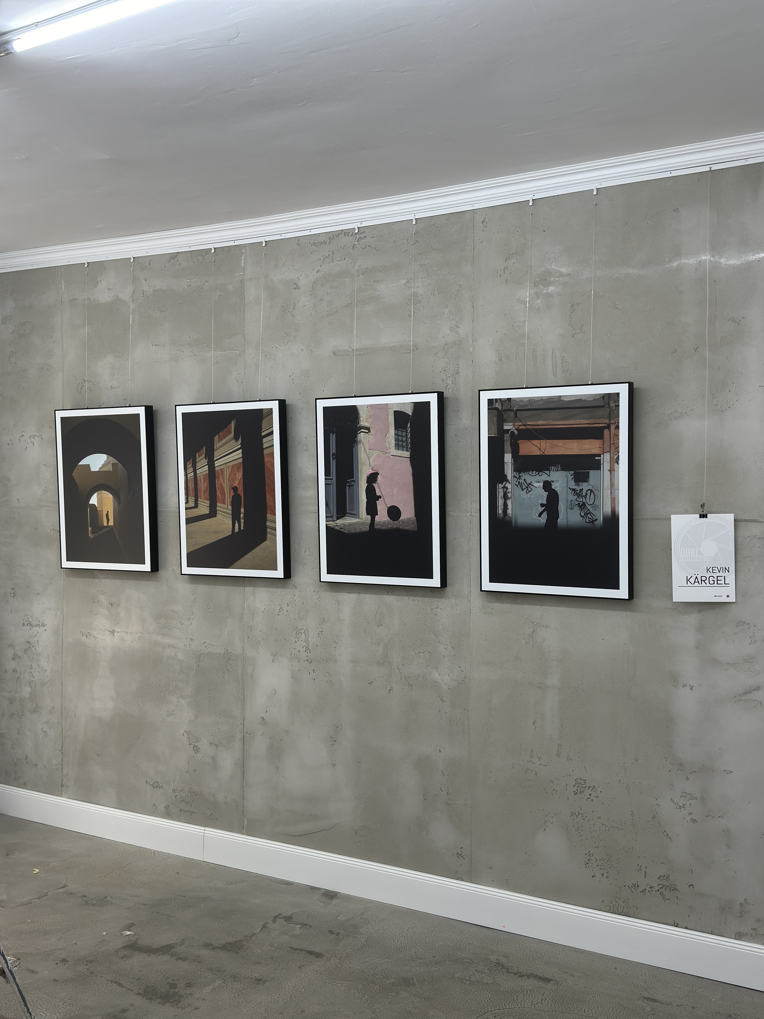 Fotografie von einer Kunstausstellung mit vier gerahmten Fotografien an einer Betonwand. Es sind Silhouetten von Menschen vor architektonischen Hintergründen zu sehen. Ein Schild mit dem Namen Kevin Kärgel ist ebenfalls sichtbar.