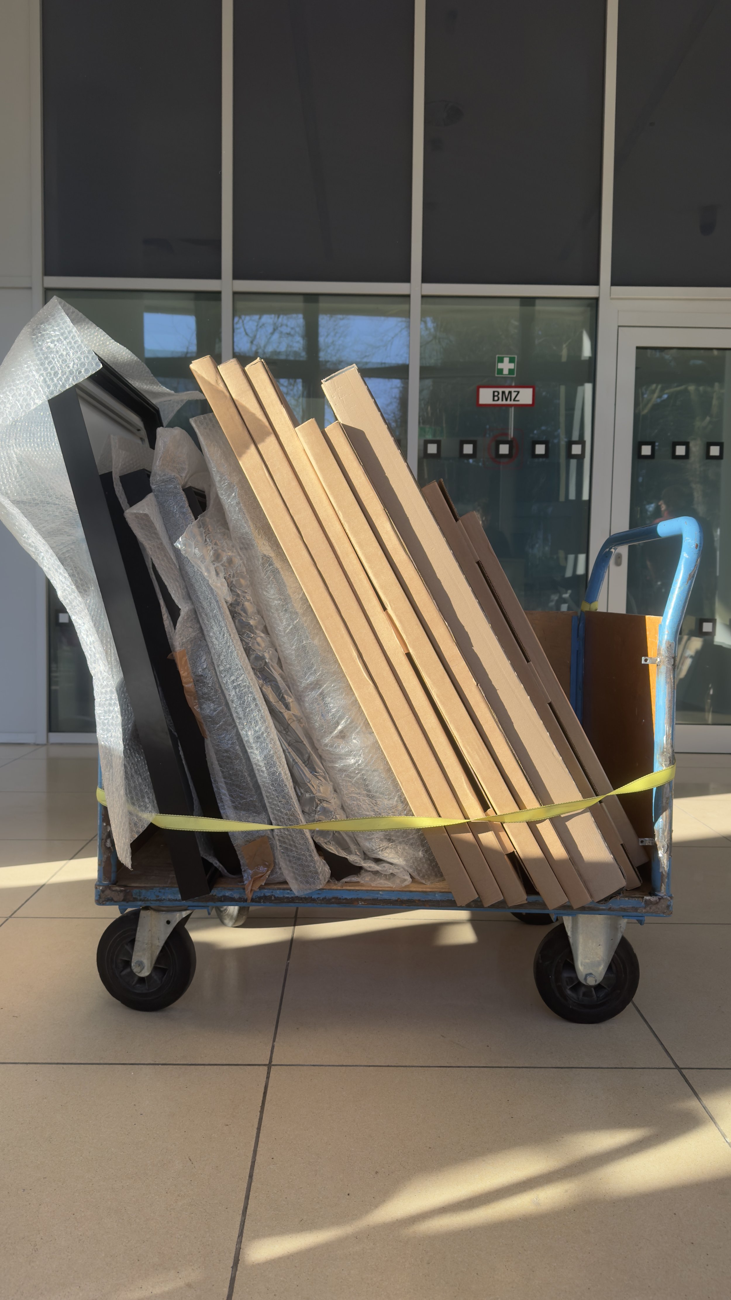 Ein Rollwagen mit diverses Verpackungsmaterialien und Kartons, vor einer Gebäudetür mit Schildern. Eyes-closed, sunlight, indoor.
