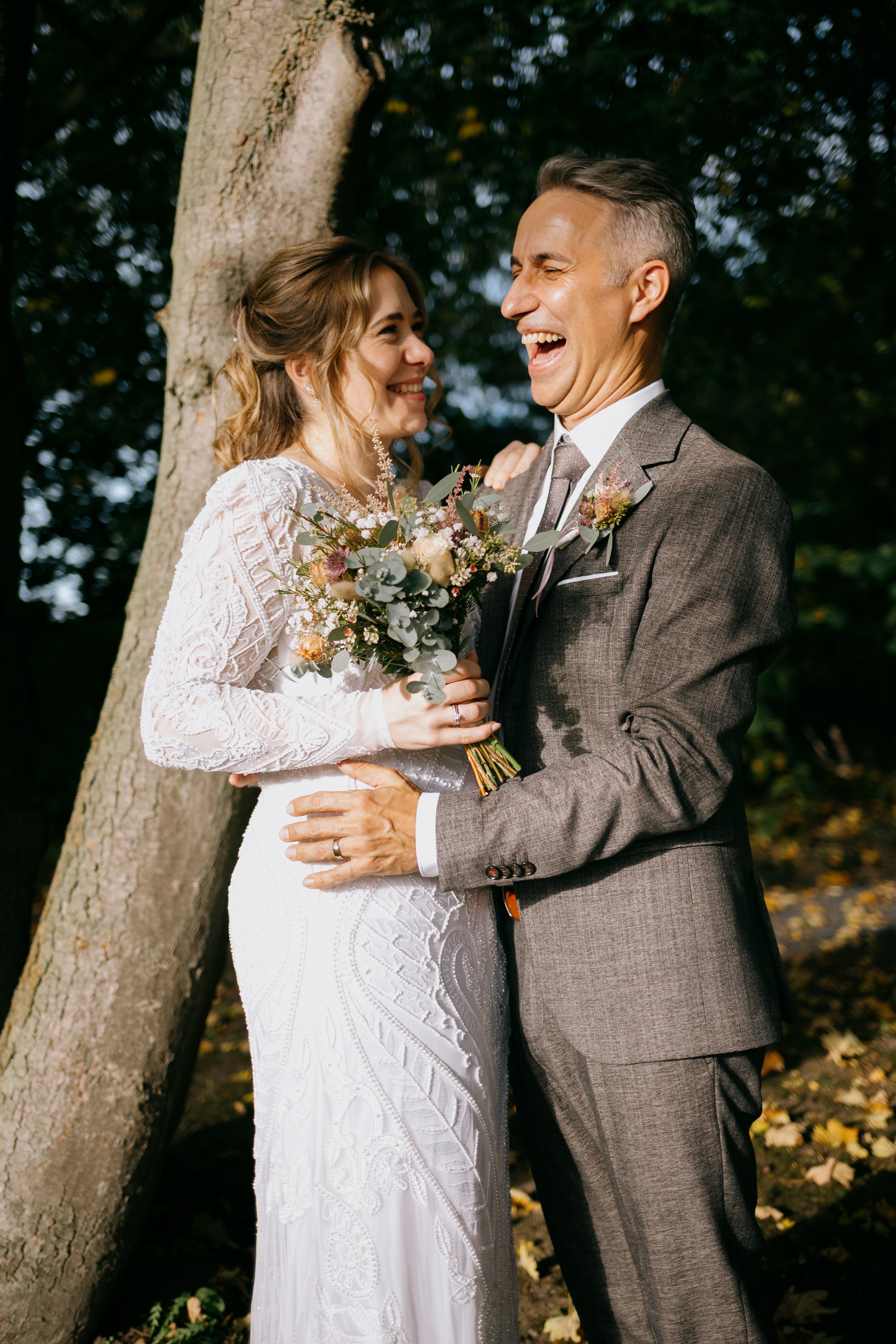 Ein lachendes Brautpaar im Wald, die Braut hält einen Blumenstrauß, sie trägt ein weißes, verzierungreiches Hochzeitskleid, der Bräutigam in einem braunen Anzug.